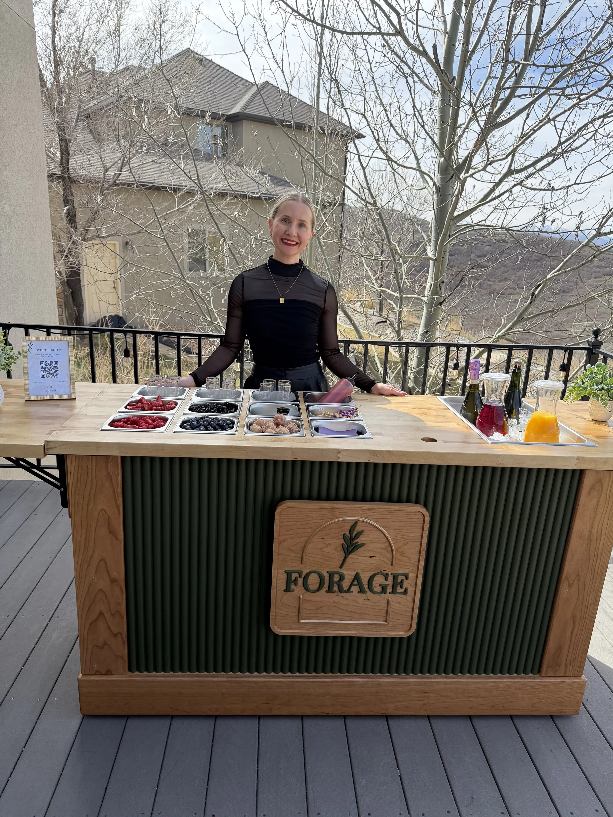 A woman behind a wooden outdoor bar with a sign that reads "FORAGE." The bar has various bowls of berries and a few bottles of juice or wine. There are trees in the background and a house with tan walls and a gray roof.