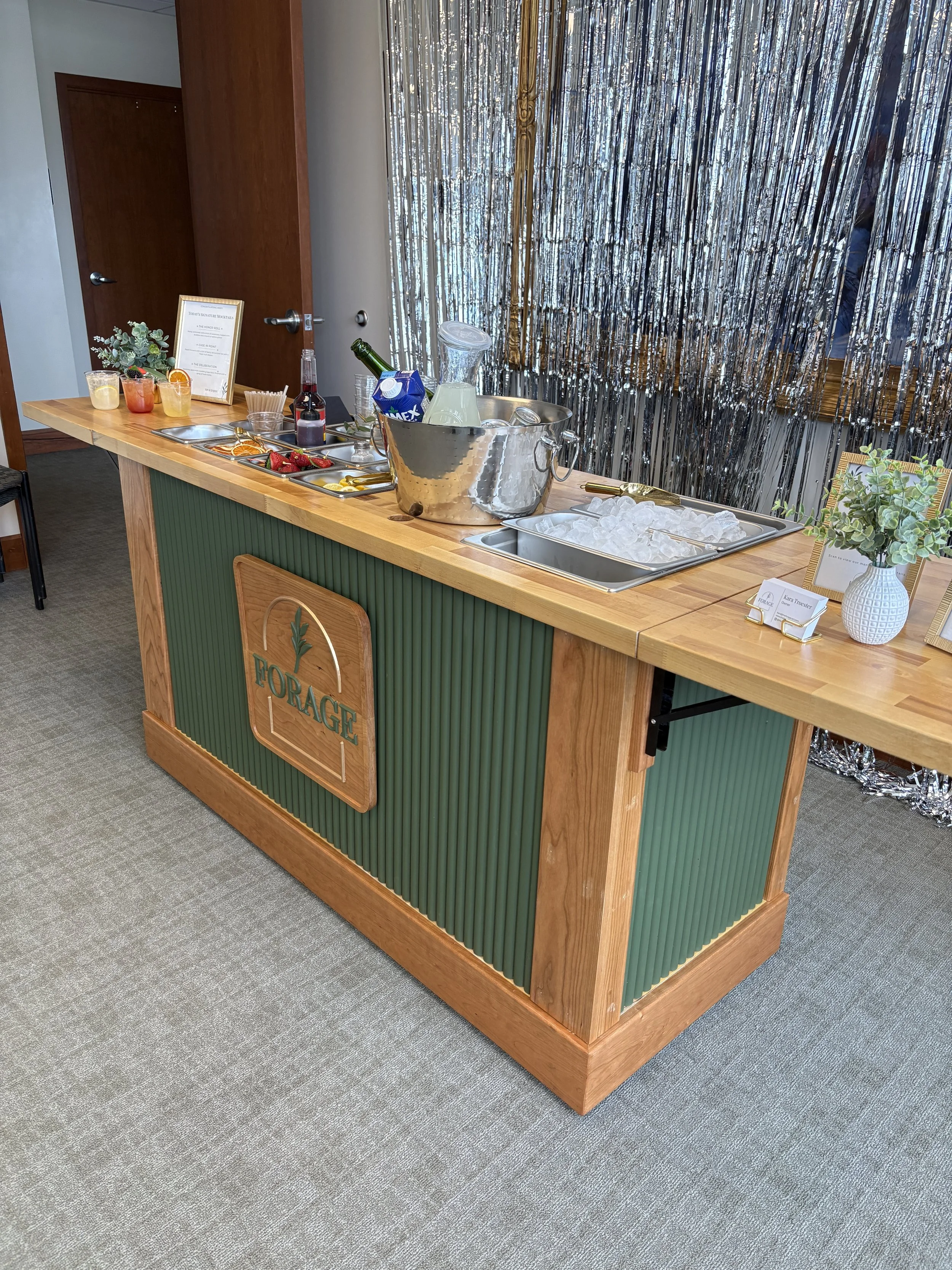 A wooden bar or countertop with a green paneling front and a sign that says "FORAGE". There are various drinks and glasses, a metal bucket with ice and bottles, small trays with strawberries and lemon slices, and a small potted plant with green leave