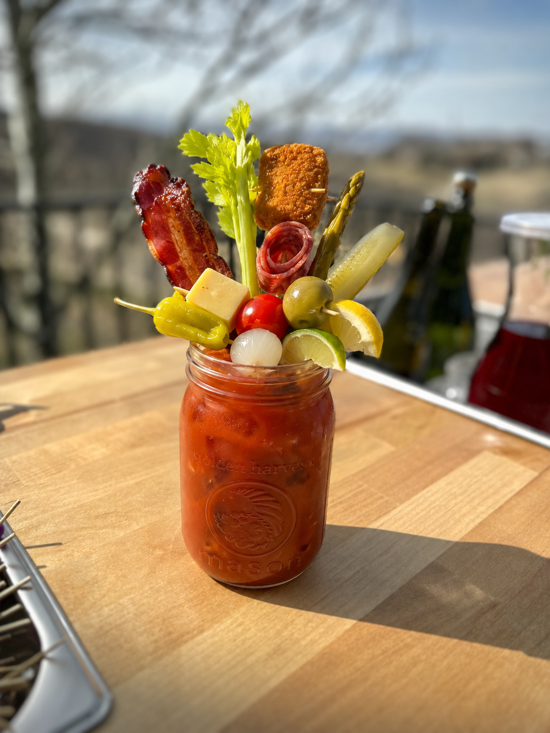 A mason jar filled with tomato juice, topped with various garnishes including bacon, celery, cheese, cherry tomatoes, pickles, lemon wedges, and olives, set on a wooden table outdoors.