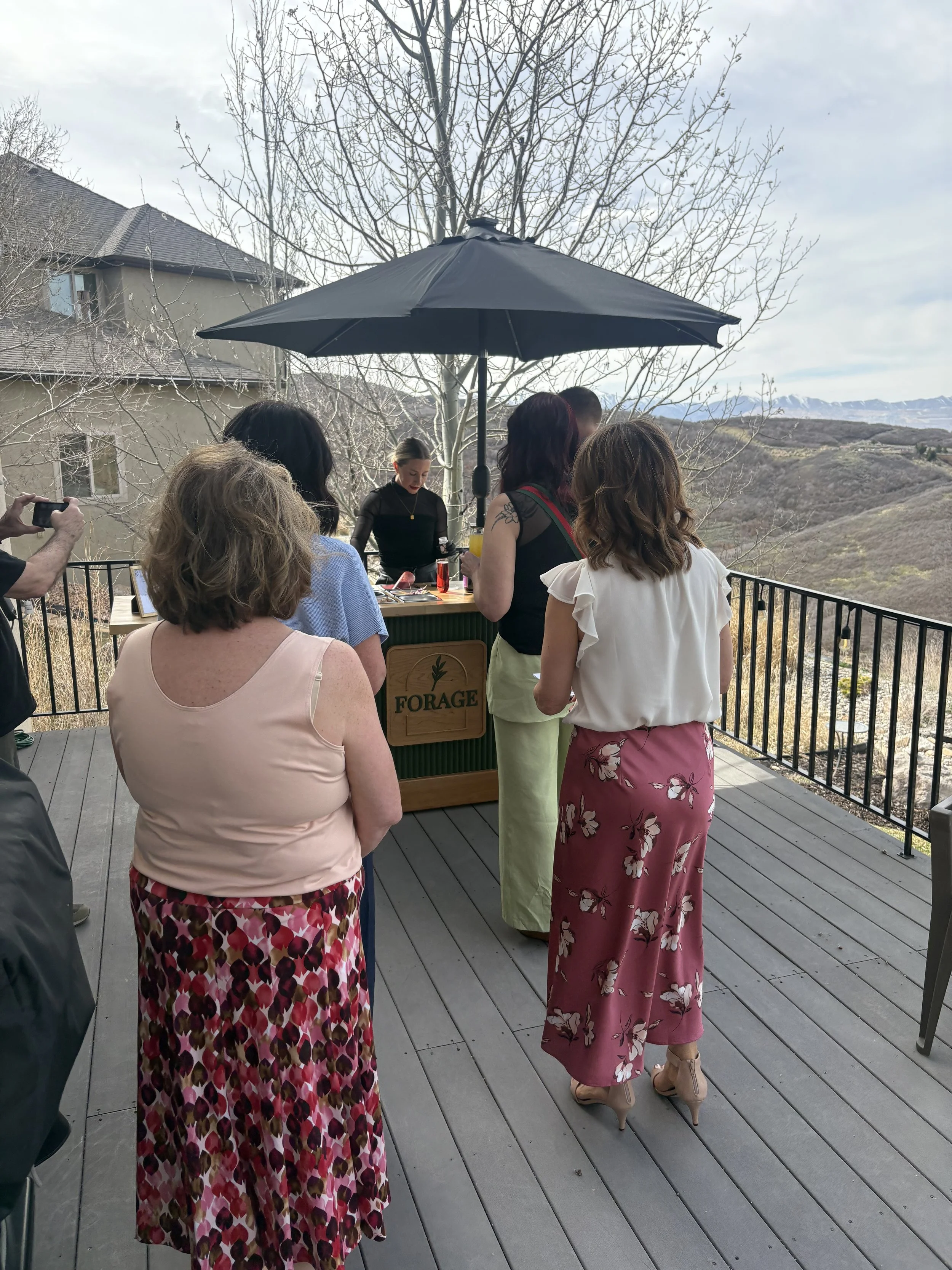 People gathered on a wooden terrace outdoors, with a black umbrella overhead. The scene shows a mountainous landscape in the background and leafless trees, suggesting early spring or late fall. The group includes women wearing casual summer clothing,