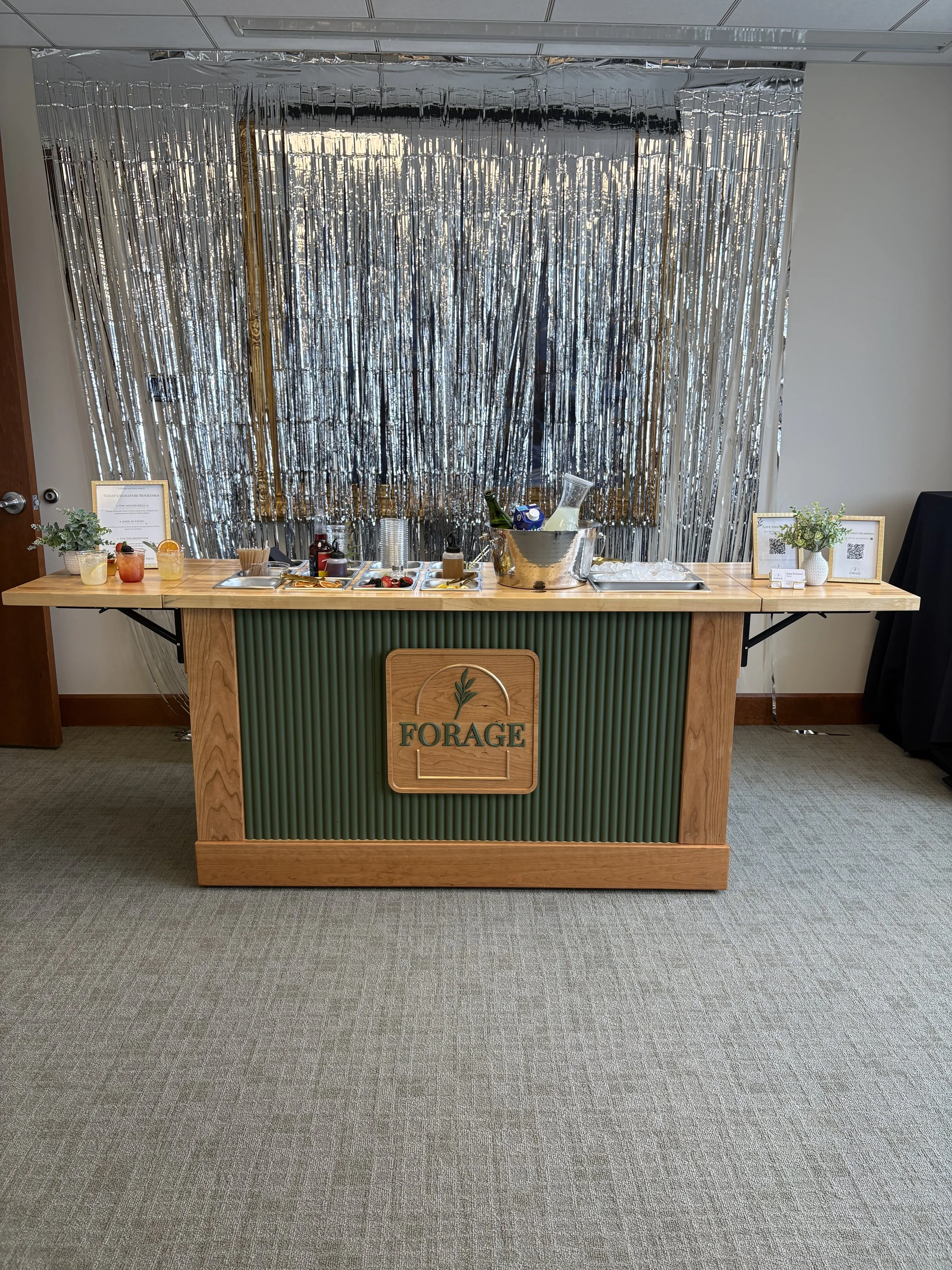 A wooden table with a green vertical slat front, a shop sign reading 'FORAGE', and a variety of drinks, snacks, and condiments, set against a metallic fringe backdrop in an indoor setting.