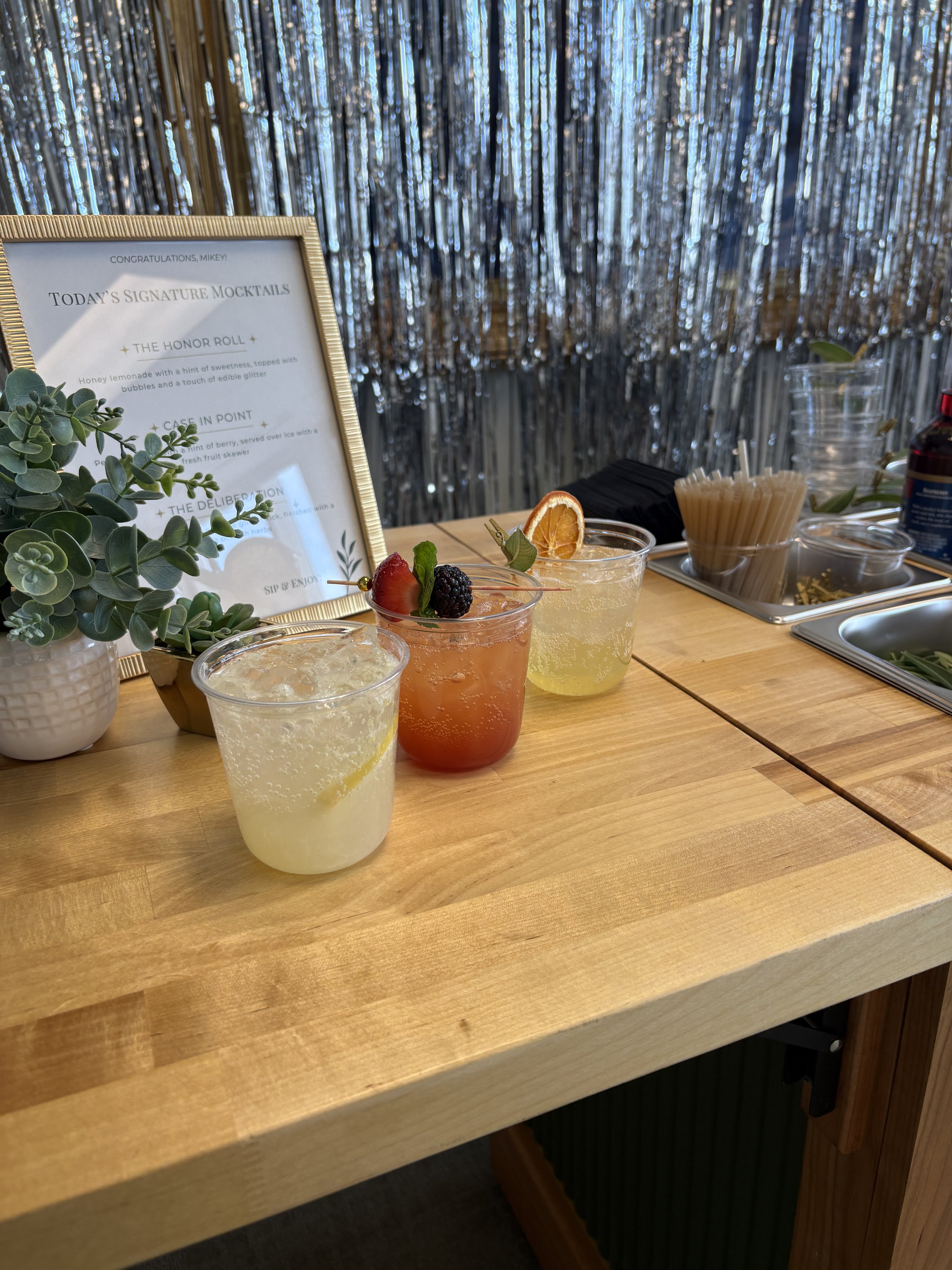 A wooden table with mixed drinks garnished with fruit, a menu stand, a potted plant, and bar accessories, with a metallic curtain backdrop.