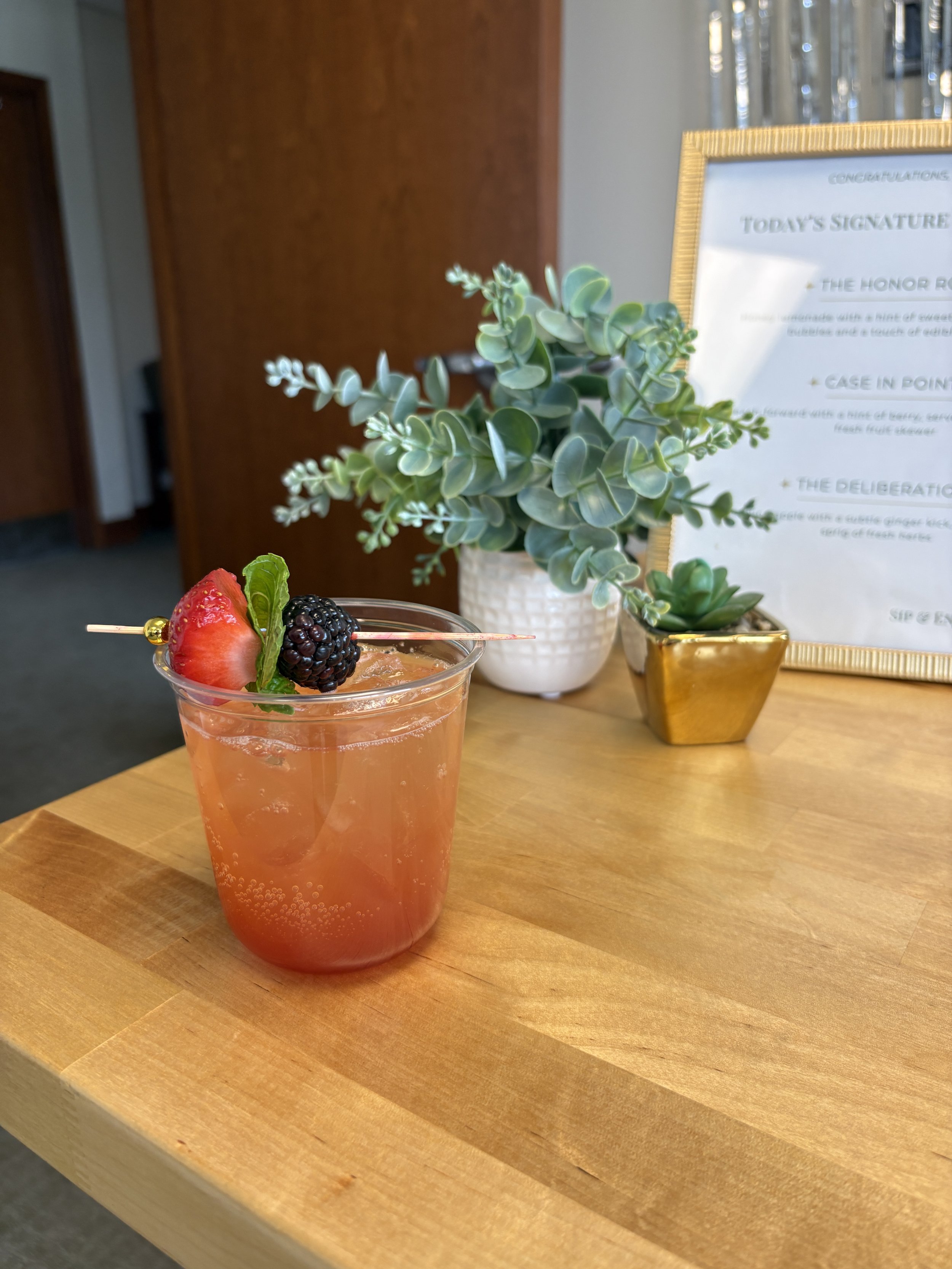 A pink cocktail garnished with a strawberry, blackberry, and a small mint leaf on top, placed on a wooden table.
