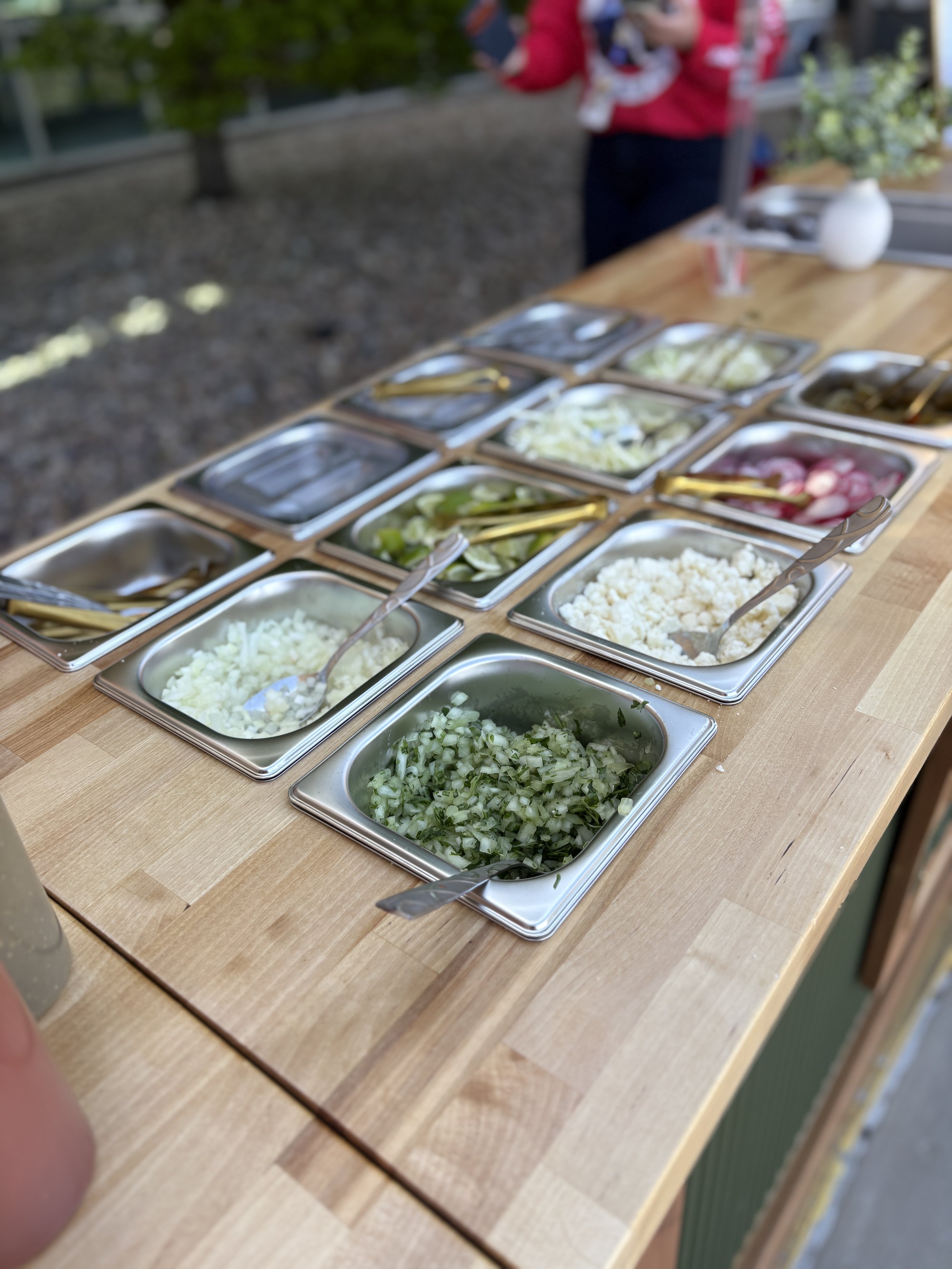 A buffet of chopped vegetables, including onions, cucumbers, and radishes, in metal trays with gold tongs on a wooden table.