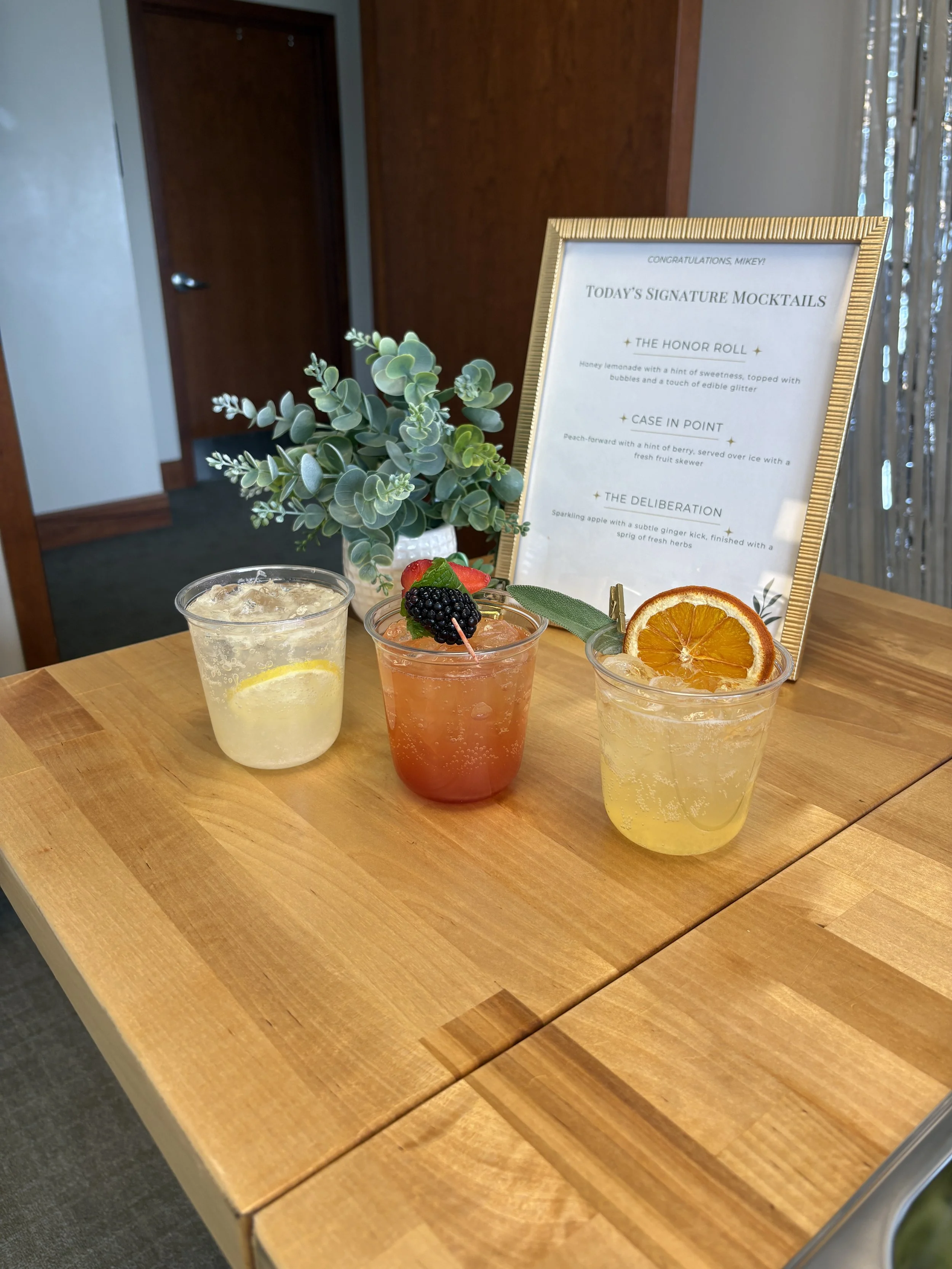Three drinks with slices of citrus and fruit garnishes on a wooden table, decorated with a green plant and a sign about signature mocktails.