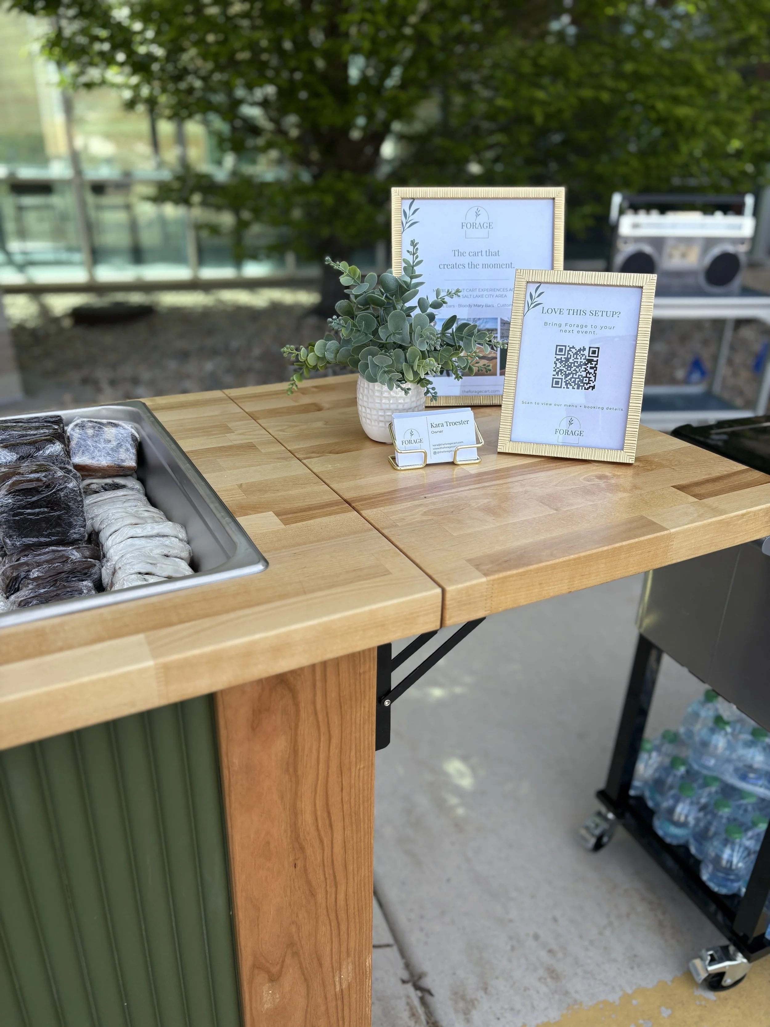 A wooden table displaying signs with information, a small potted plant, and a tray of wrapped cookies at an outdoor event. In the background, there is a blurred view of a tree and a vintage radio.
