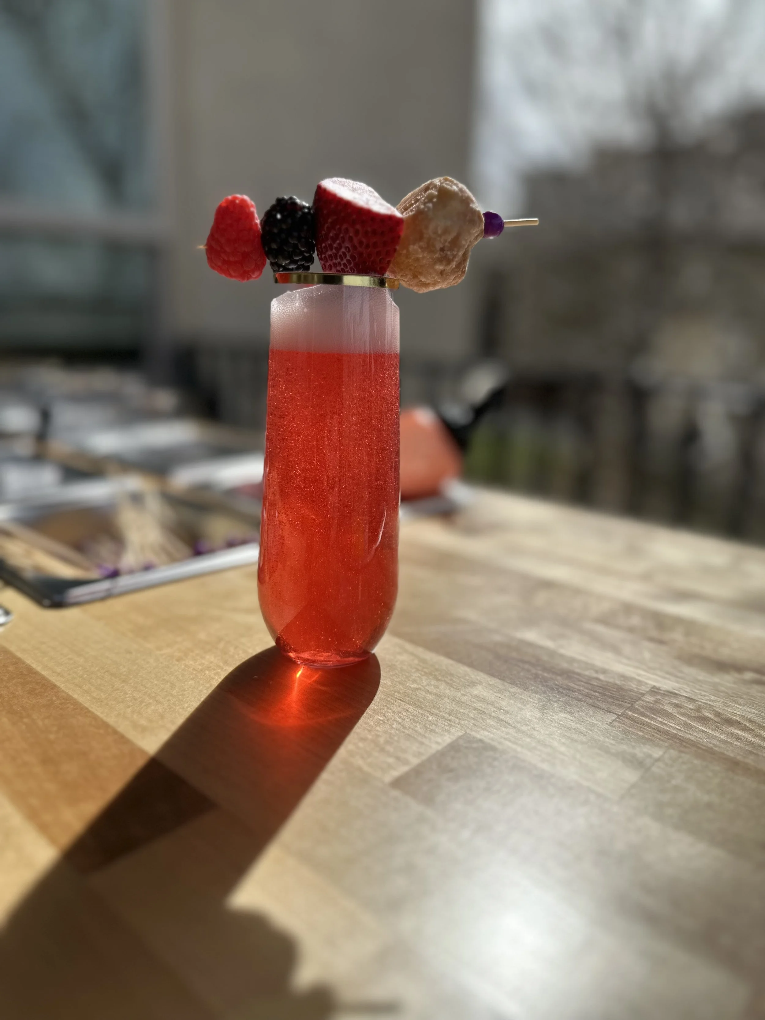 A red cocktail with a slice of strawberry, blackberry, and a piece of peach on a skewer on top, casting a shadow on a wooden surface.