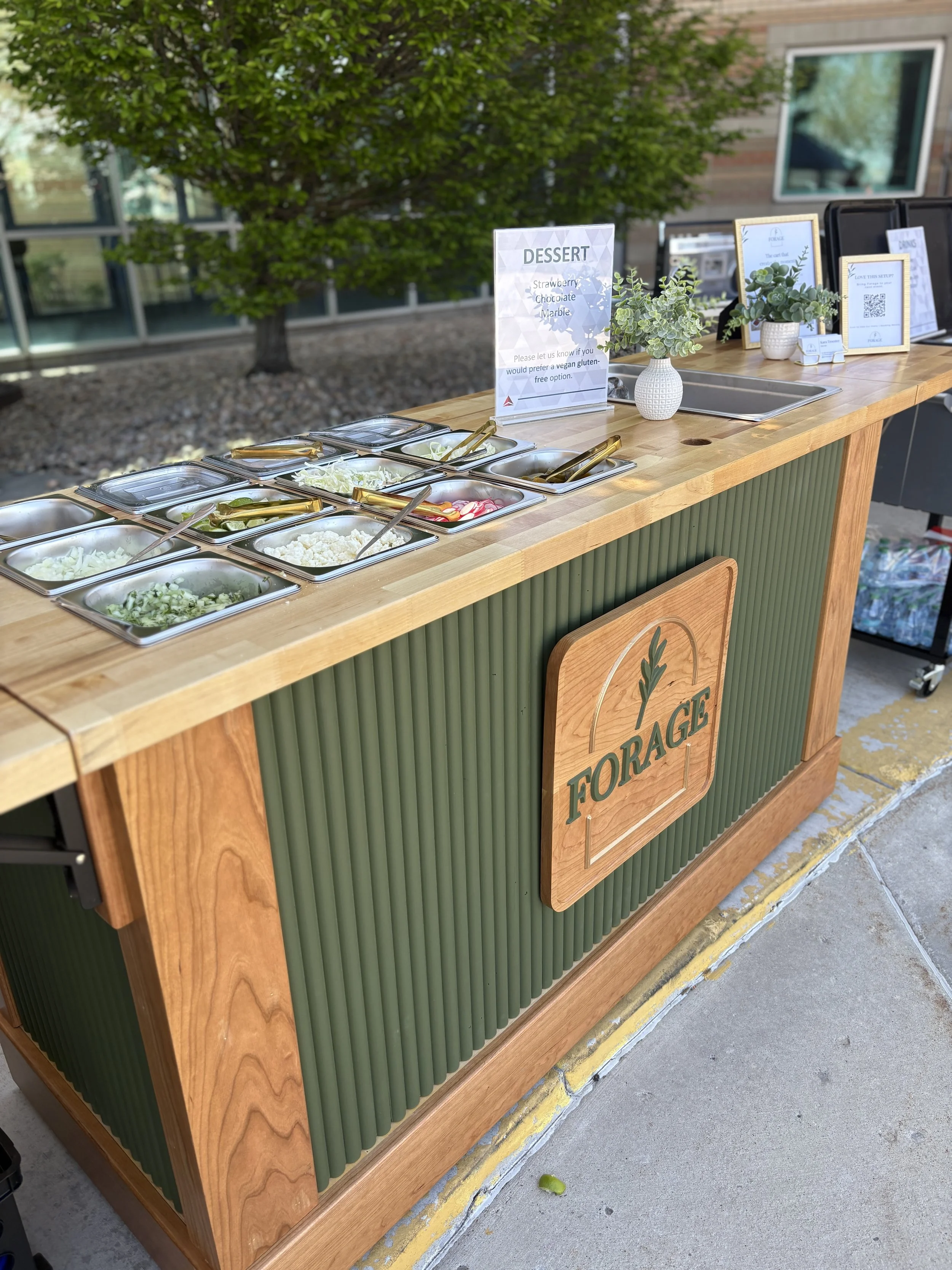 Outdoor food stand with a sign labeled 'DESSERT' listing strawberry, chocolate, and marble options. The stand has small containers of chopped vegetables or toppings, a decorative potted plant, and informational signs, set on a sidewalk with a tree an