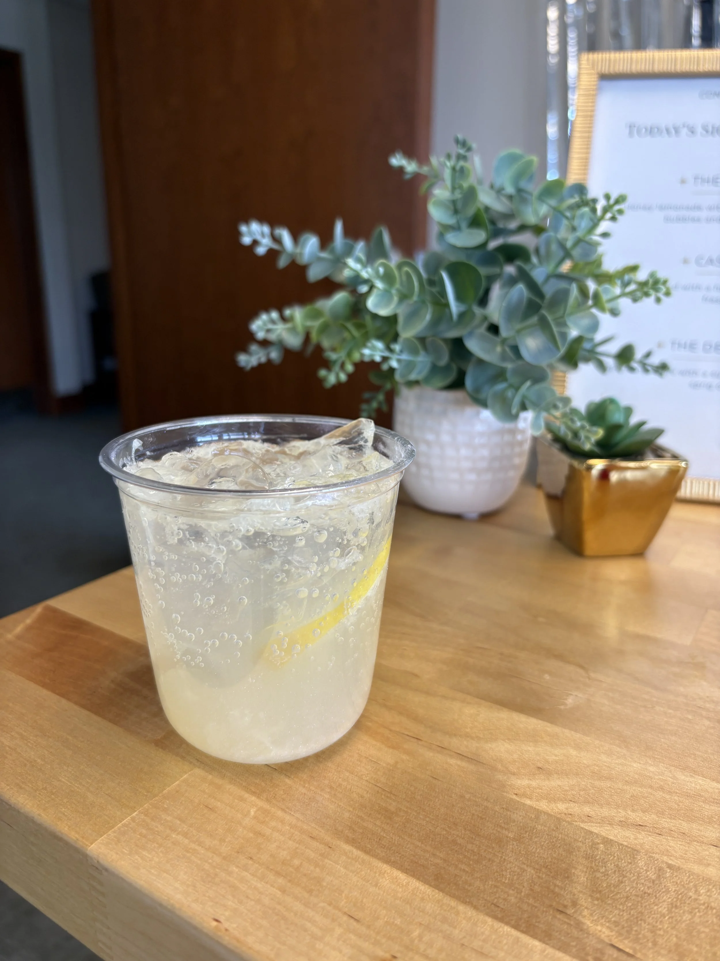 A glass of lemonade with ice and lemon slices on a wooden table in a cafe. In the background, there's a potted plant with green leaves and a framed menu.