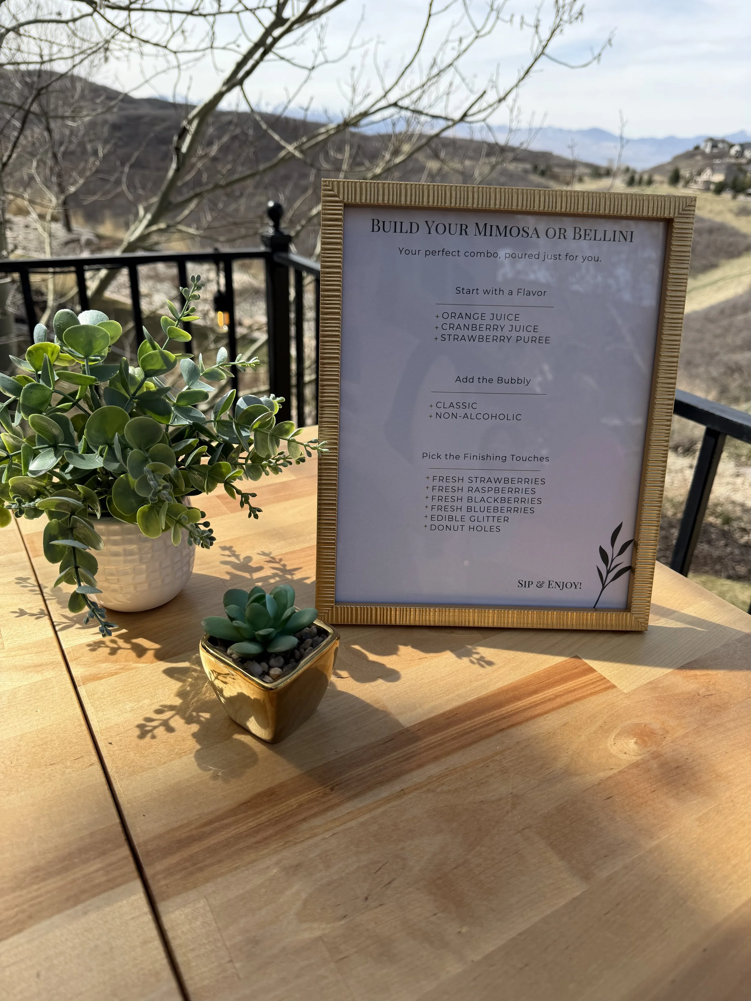 A menu card on a wooden table with potted succulents and a decorative plant. The menu provides instructions for building a mimosa or bellini, including flavor options, bubbly choices, and finishing touches.