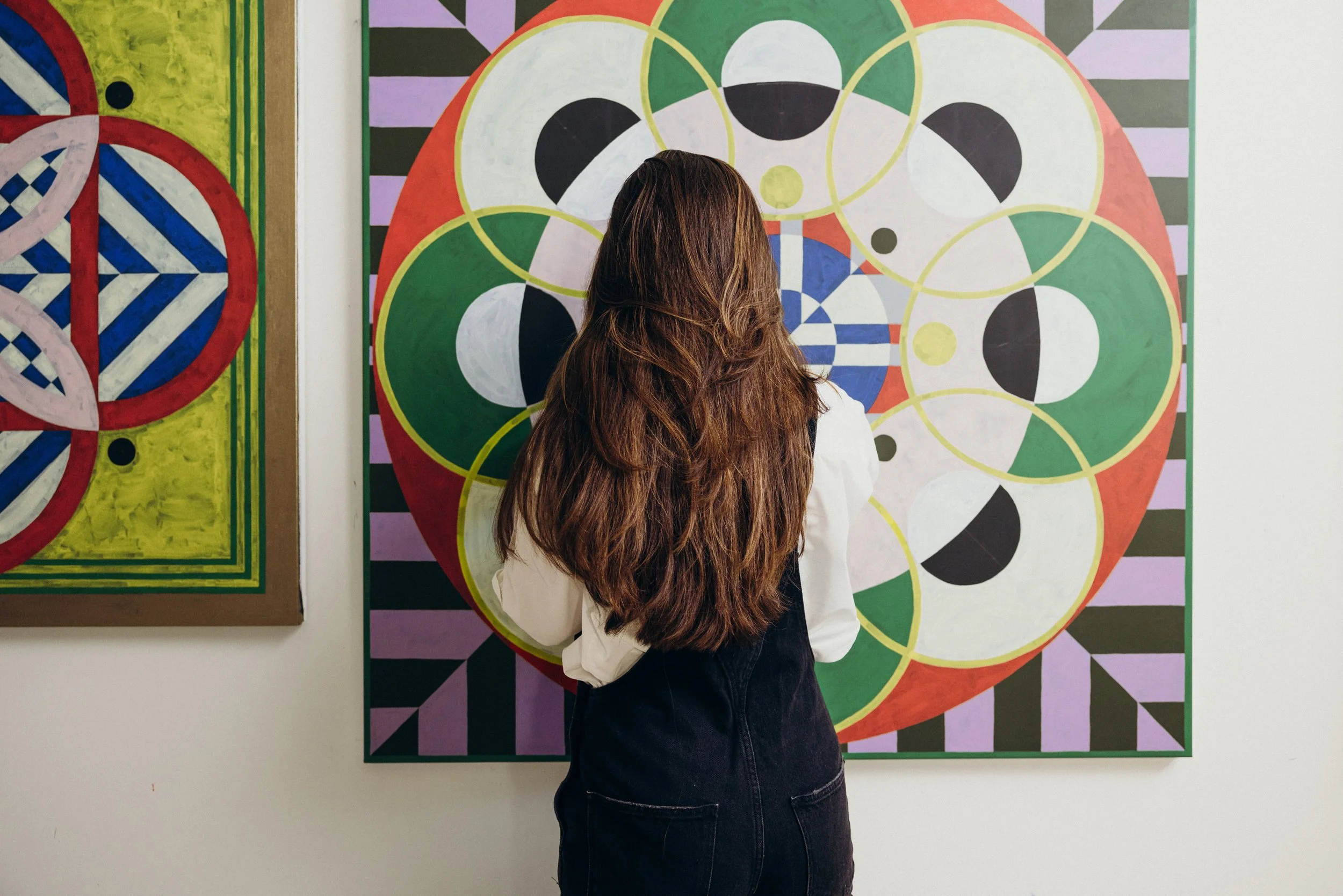 Kate wearing a white shirt and black overalls stands with her back to the camera, painting a colourful abstract painting in her studio. Photo by Nick George