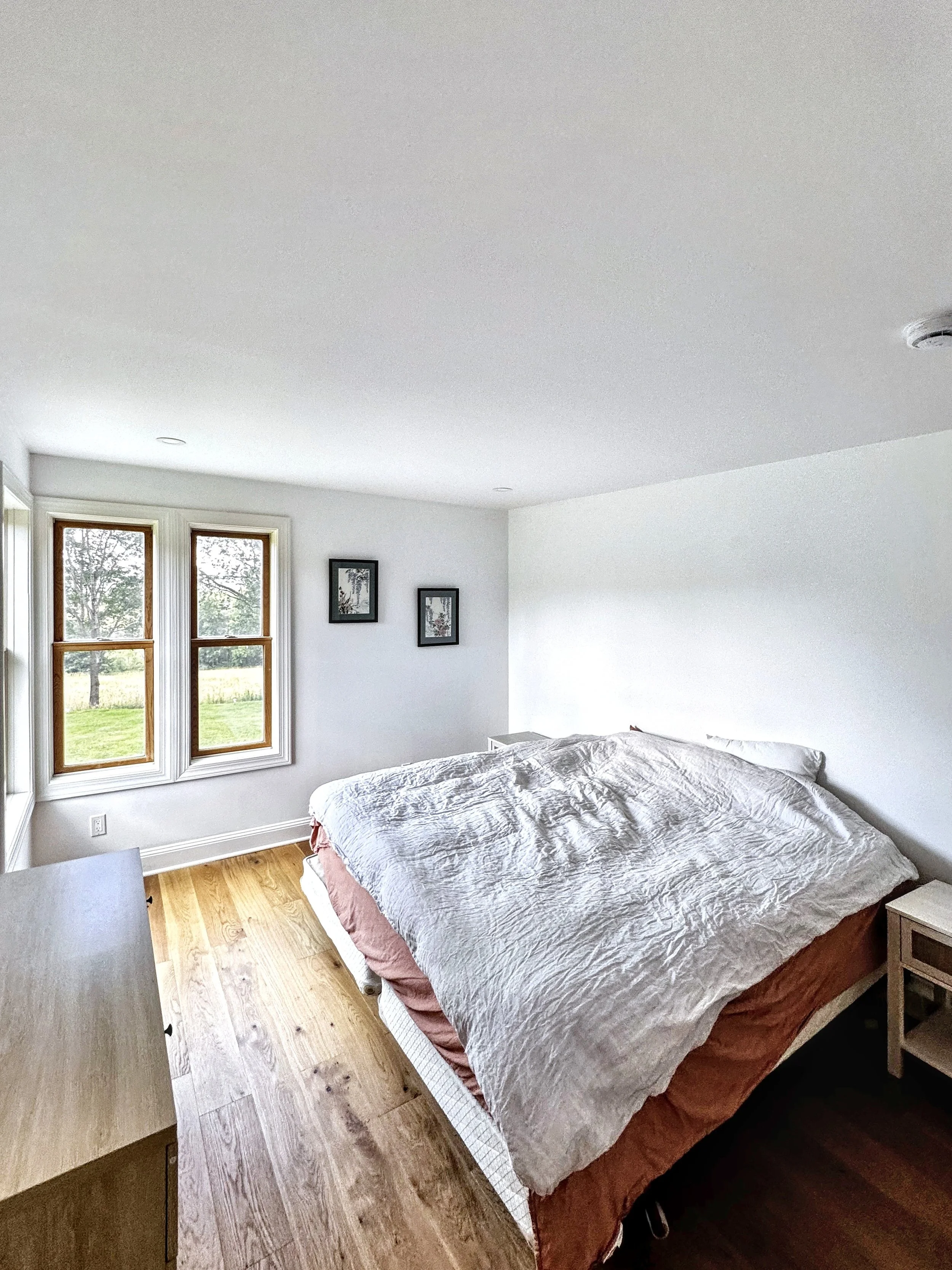 A bedroom with white walls, hardwood floors, a large bed with white and brown bedding, two windows with wooden frames showing a grassy outdoor scene, and three black picture frames on the wall.