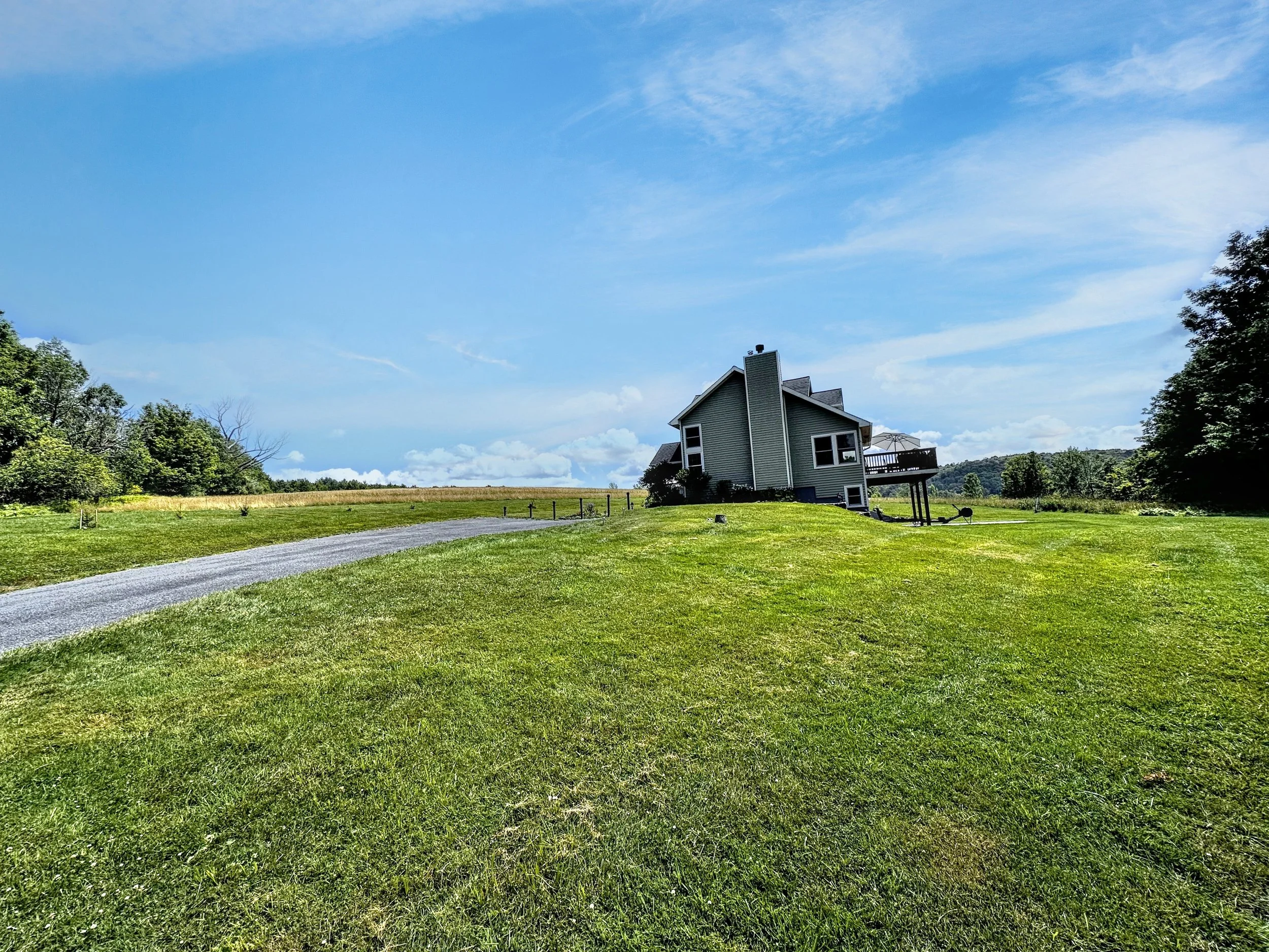 A house on a grassy hill under a blue sky with some clouds, surrounded by trees and open fields.