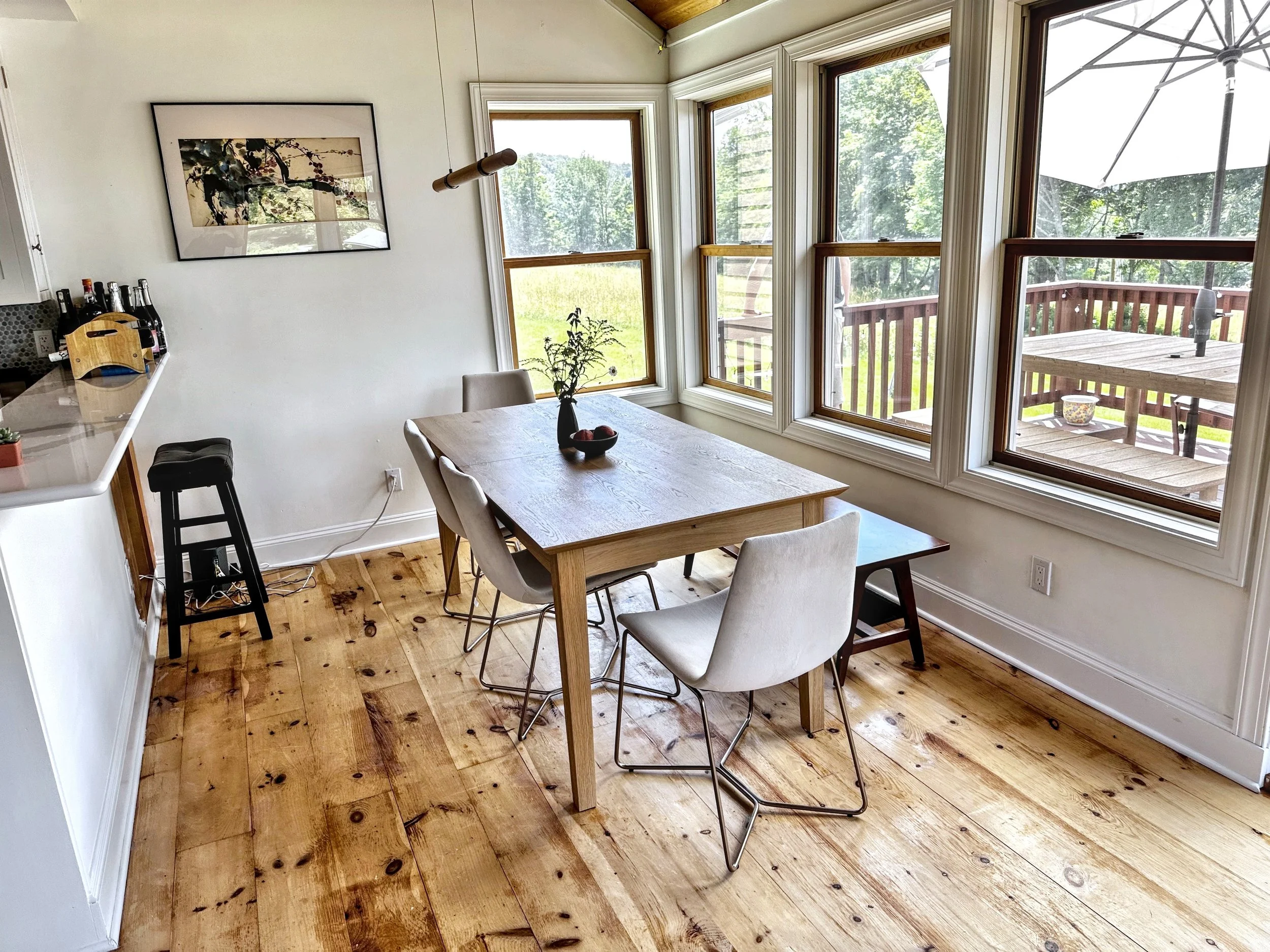 Bright dining area with a wooden table, four white chairs, and large windows overlooking a deck and green trees outside.