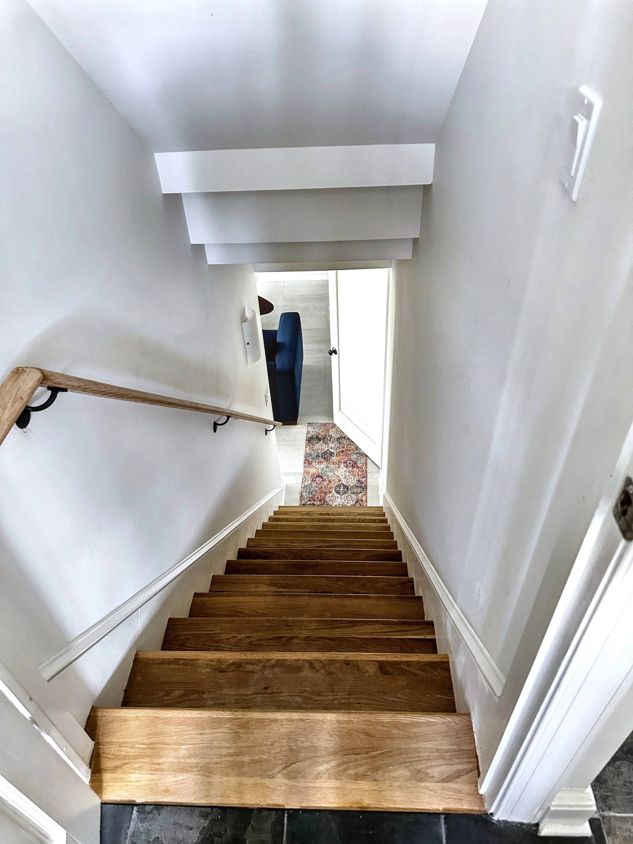 View of a staircase looking downward towards a door with a small rug in front of it, with a seating area partly visible at the bottom.
