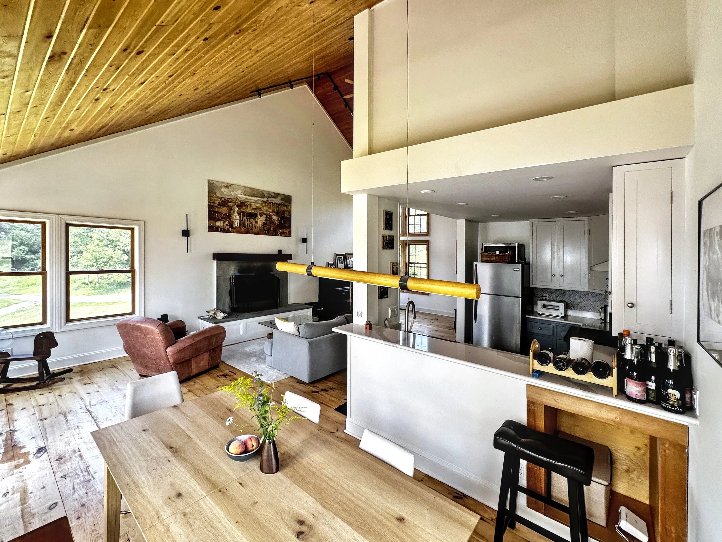 Open-concept living room and kitchen with wooden floors, white walls, a vaulted wooden ceiling, large windows, and modern furniture including a gray sofa, a pink armchair, and a wooden dining table with chairs.