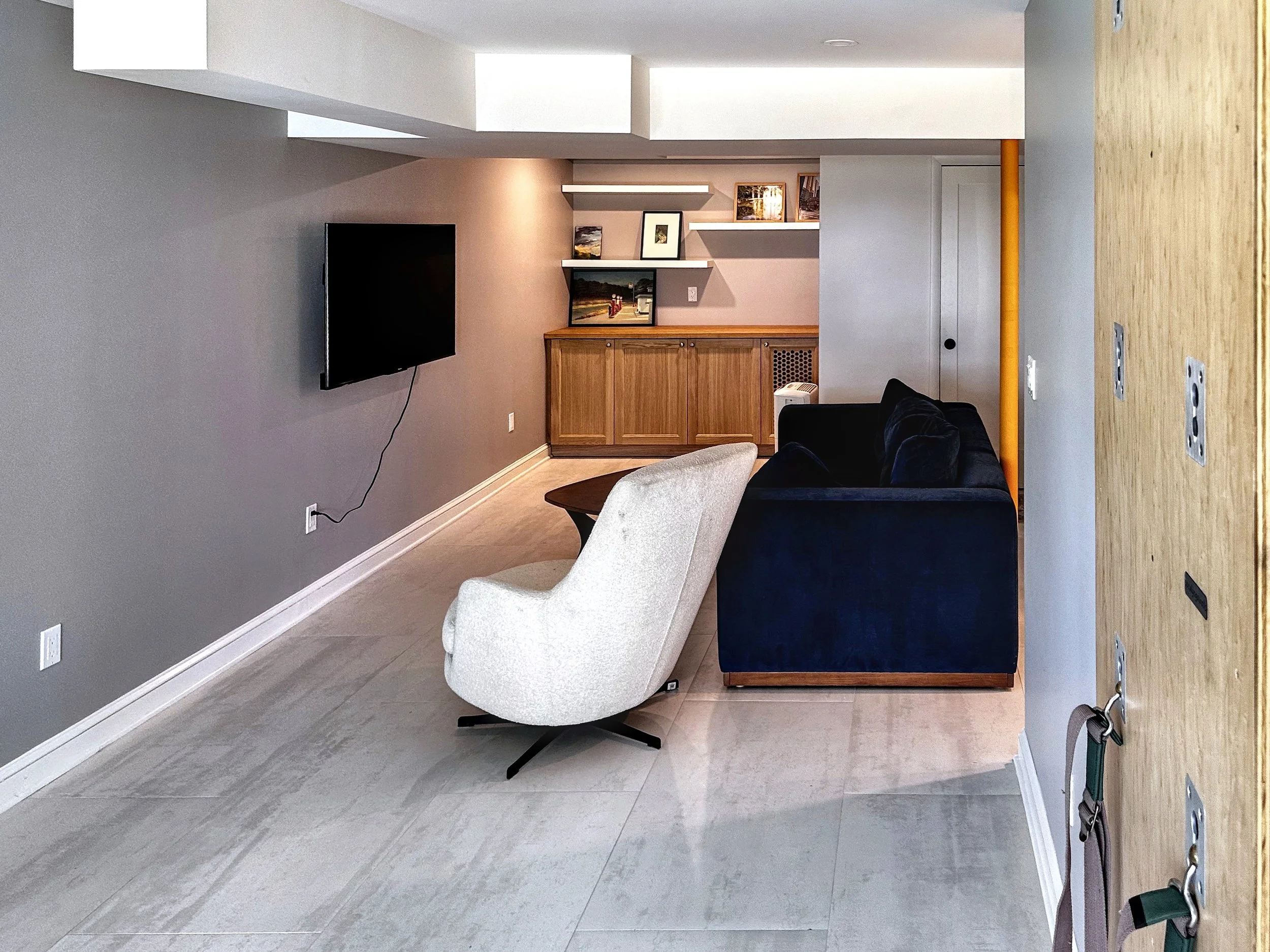 Modern living room with a wall-mounted TV, a beige swivel chair, a navy sofa, a wooden cabinet, and open shelves with framed pictures. The room has gray walls, light-colored tiled flooring, and a wooden panel with electrical outlets and hooks on the