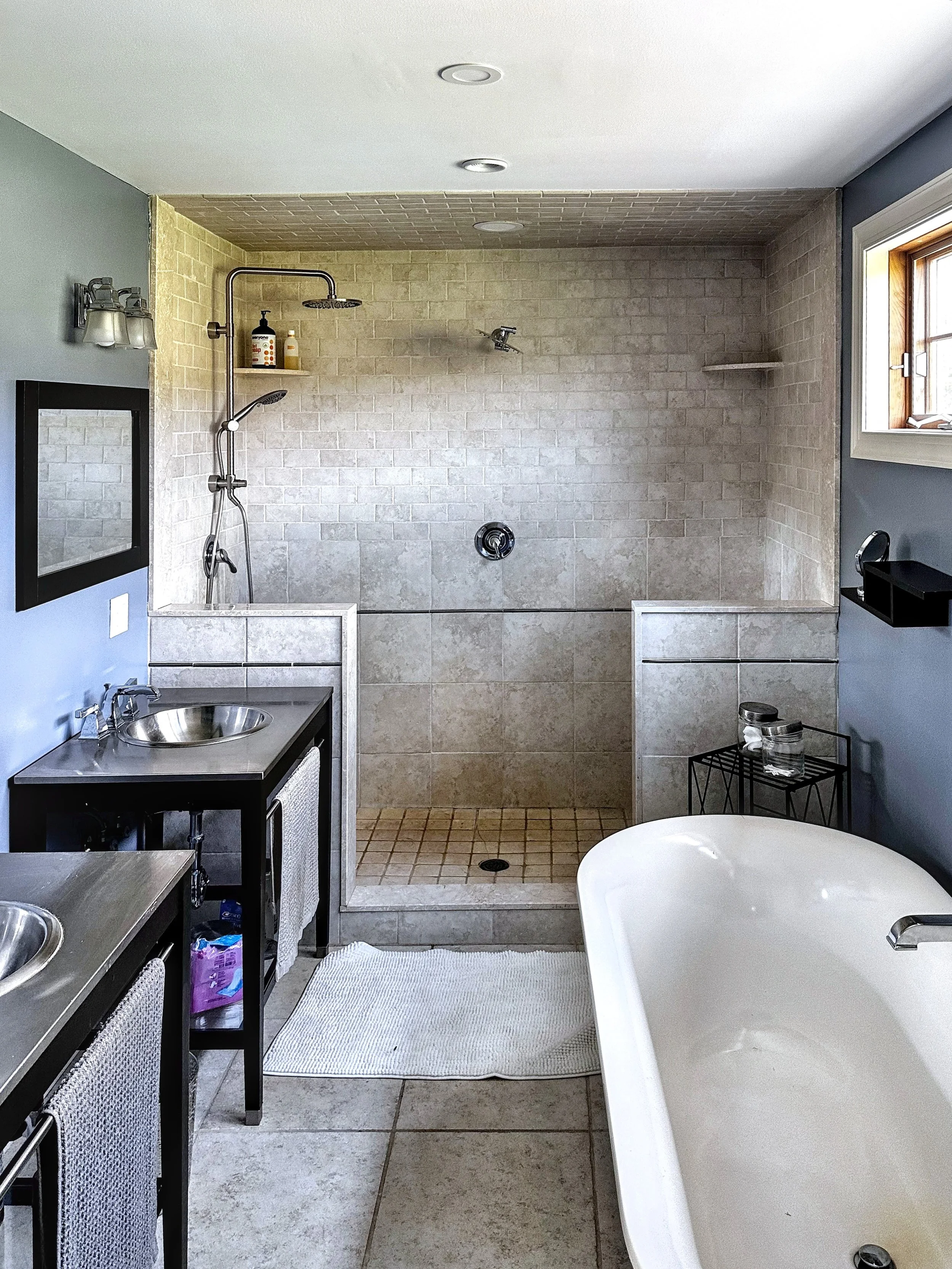 Bathroom with a large walk-in shower, a white bathtub, a double vanity with two sinks, a mirror, and a small window.