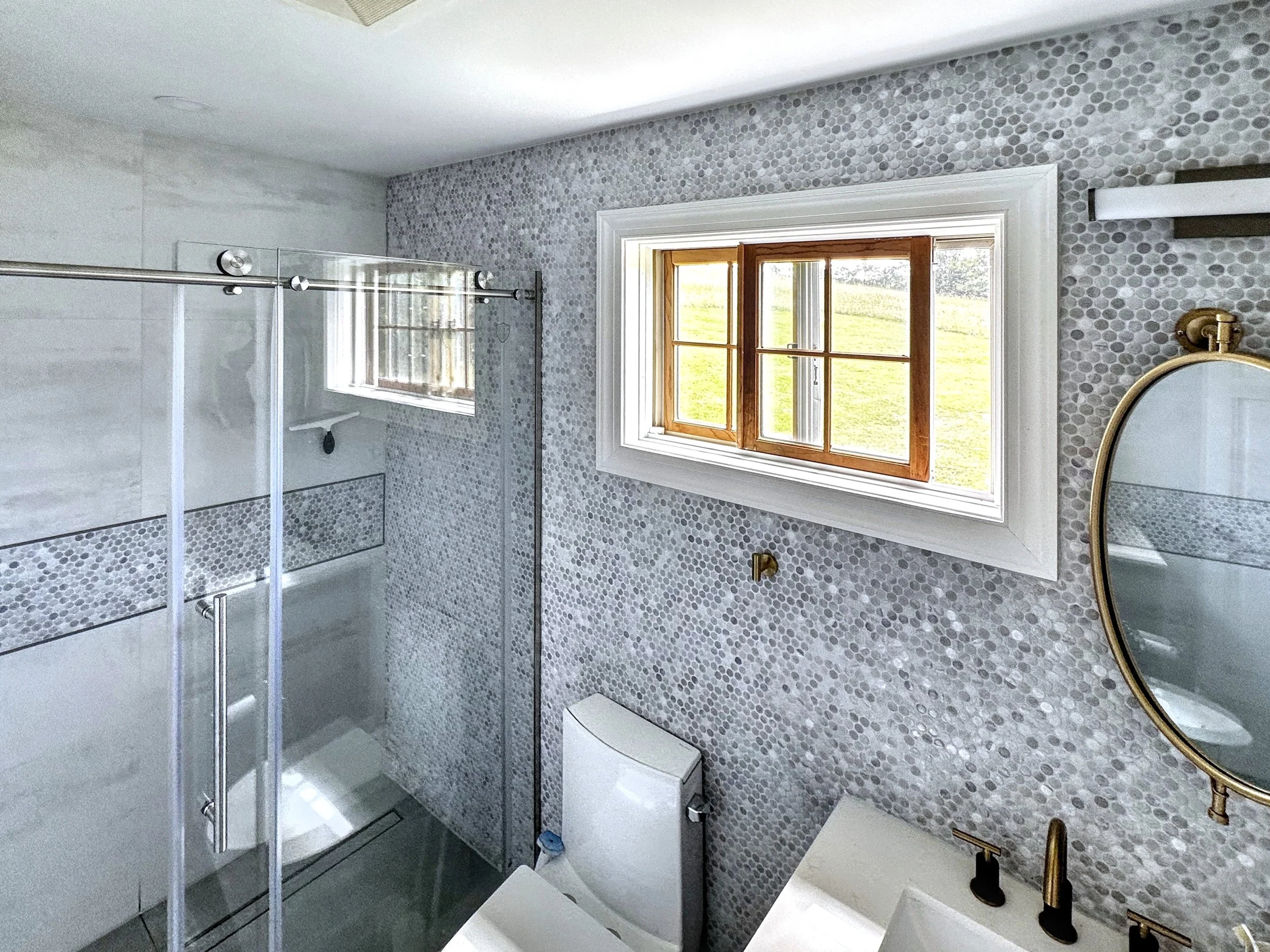 Modern bathroom with a mosaic tile wall, window with wooden frames, over a toilet, and a vanity with an oval mirror and gold fixtures.