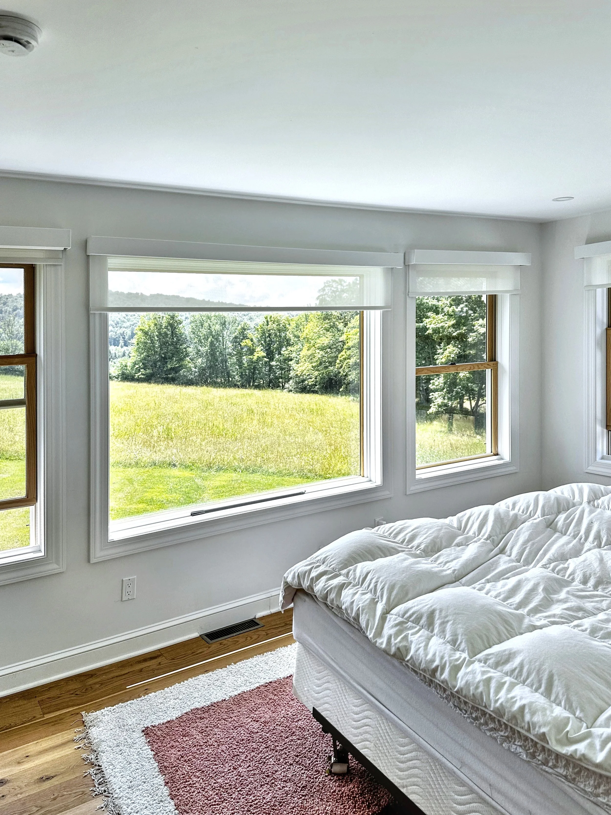 A bright bedroom with a large window overlooking a green field and trees, a white bed with a quilt, and a small rug on a wooden floor.