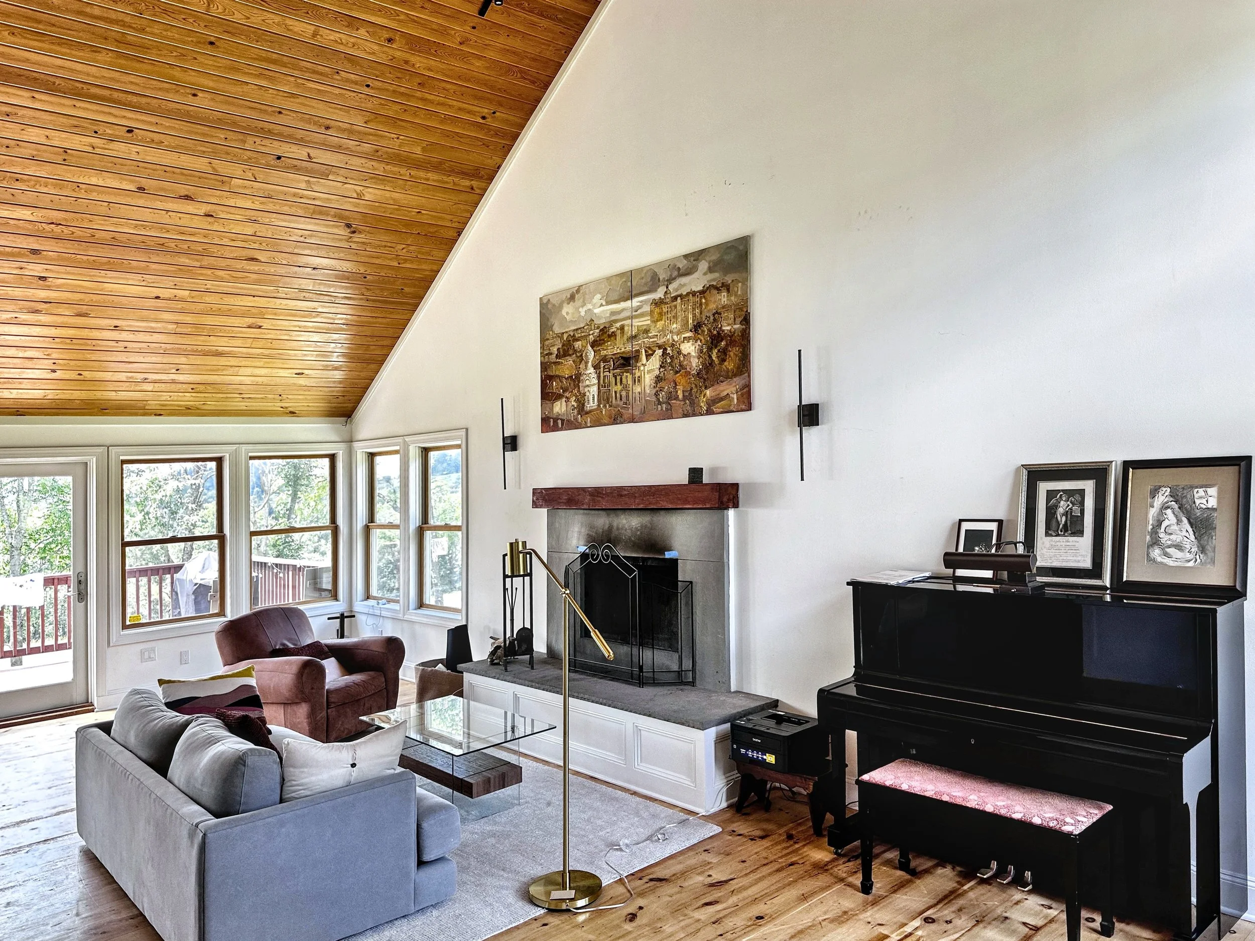 Living room with a gray couch, a maroon armchair, and a glass coffee table. Fireplace with artwork above, a black upright piano with framed pictures on top, and large windows with a view of trees outside.