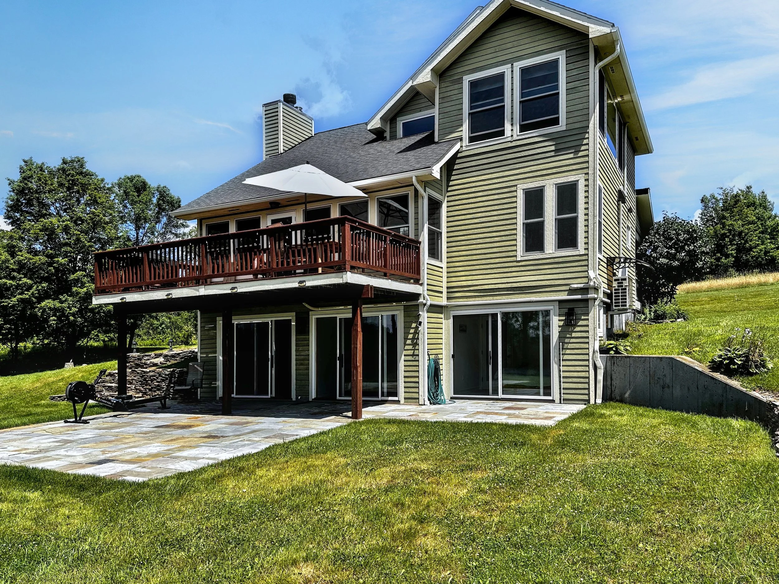 A multi-story house with beige siding, large windows, a screened-in porch, and a deck with outdoor furniture and an umbrella. The backyard has a stone patio and green grass garden with trees in the background.