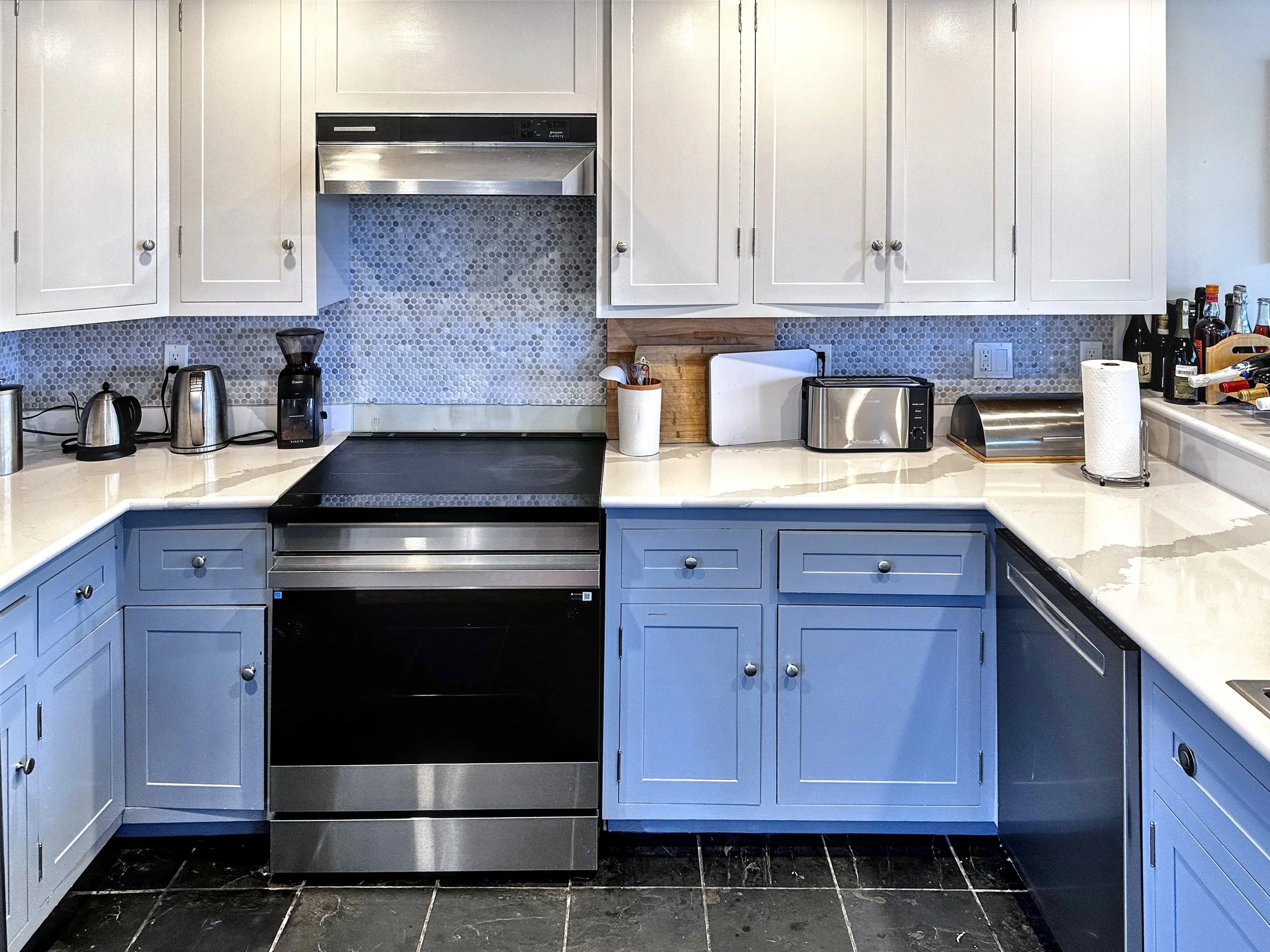 Modern kitchen with white upper cabinets, blue lower cabinets, black and stainless steel appliances including an oven, toaster, and coffee maker. The countertops are white, and there are various kitchen items like paper towel, bottles, and small appl
