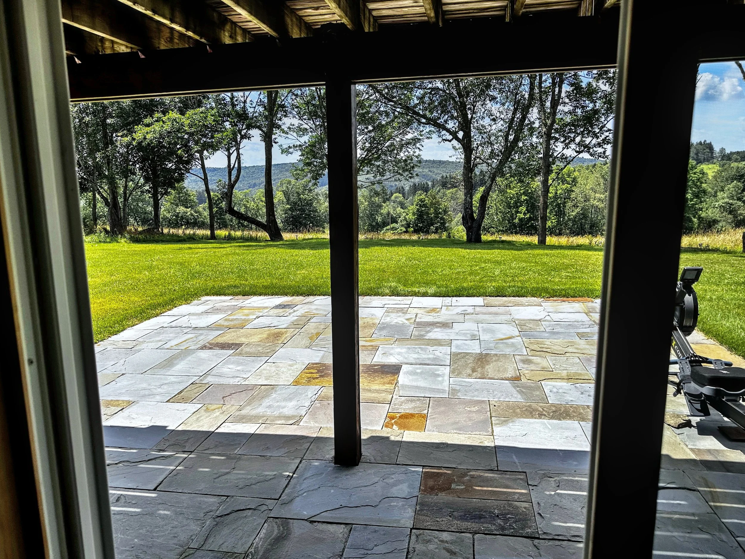 View from inside a house or patio looking out through a sliding glass door, showing a stone patio, green lawn, trees, and a distant hills or mountains under a blue sky with some clouds.