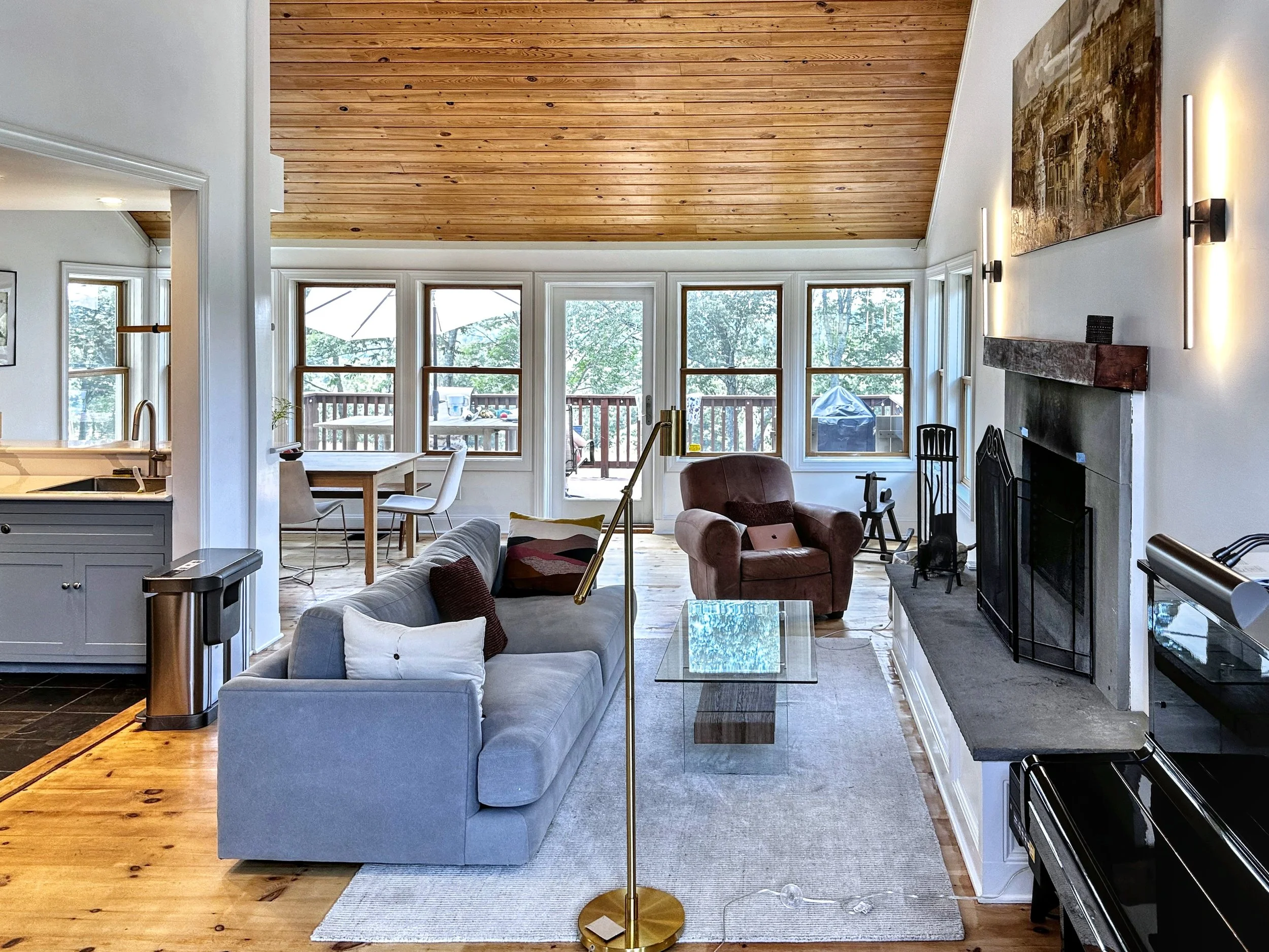 Living room with a high wood-paneled ceiling, white walls, a large window wall, and a gray couch with colorful pillows. There's a brown armchair near the fireplace, which has a wooden mantle and black fireplace tools. A glass coffee table is in front