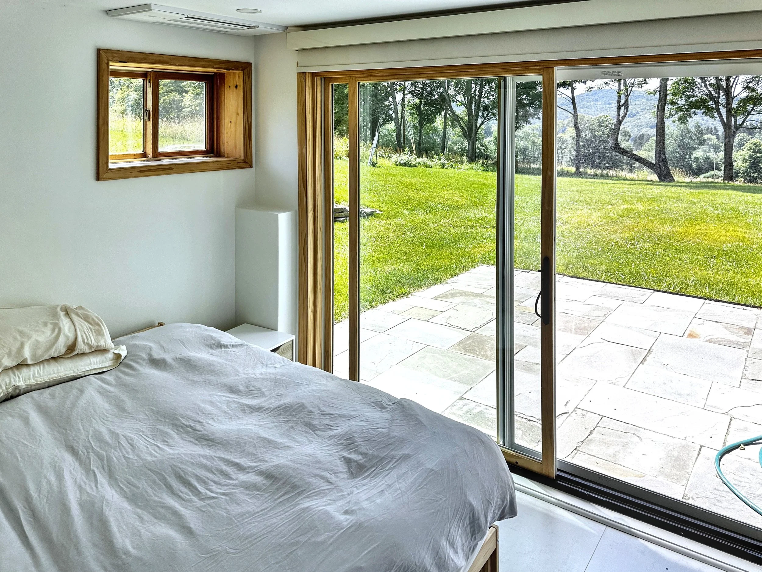 A bedroom with a bed, a window, and a sliding glass door leading to an outdoor patio with a lush green yard and trees.