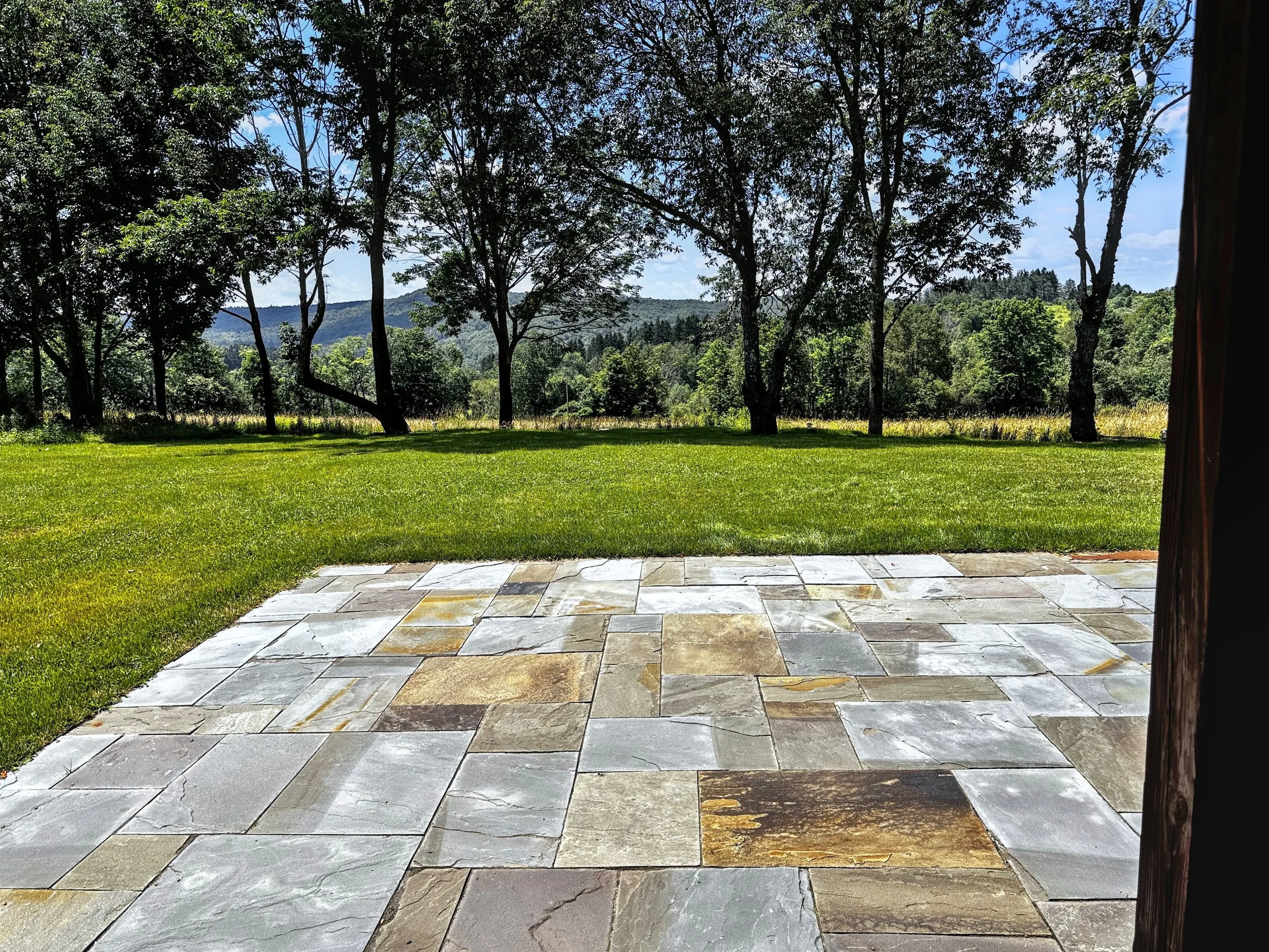 A view from a patio with a stone floor, overlooking a green lawn and forested hills under a blue sky with some clouds.