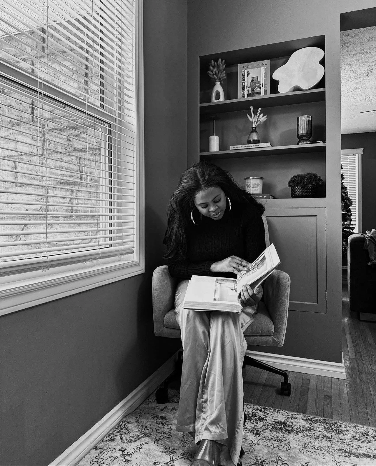 A woman sitting in a chair, reading a large book, in a cozy room with a window and a bookshelf.