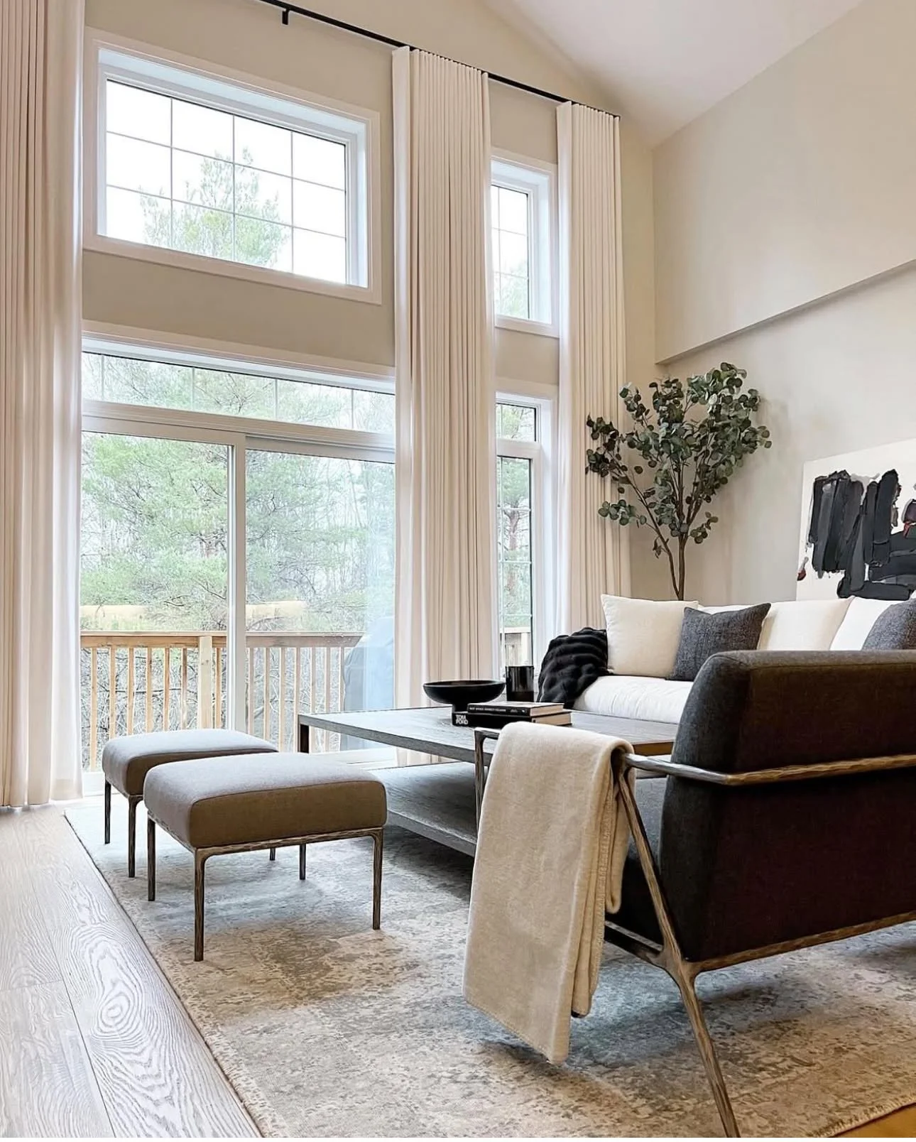 Living room with large windows, white curtains, a black and white sofa, a coffee table, two upholstered ottomans, a potted plant, and abstract artwork.
