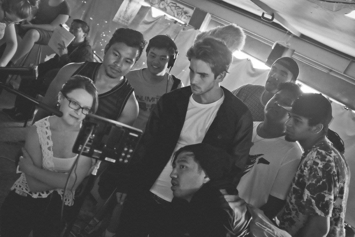 A group of eight young people, including men and women, gathered around a camera in a room with other people sitting in the background. The scene appears to be a film or photography set.