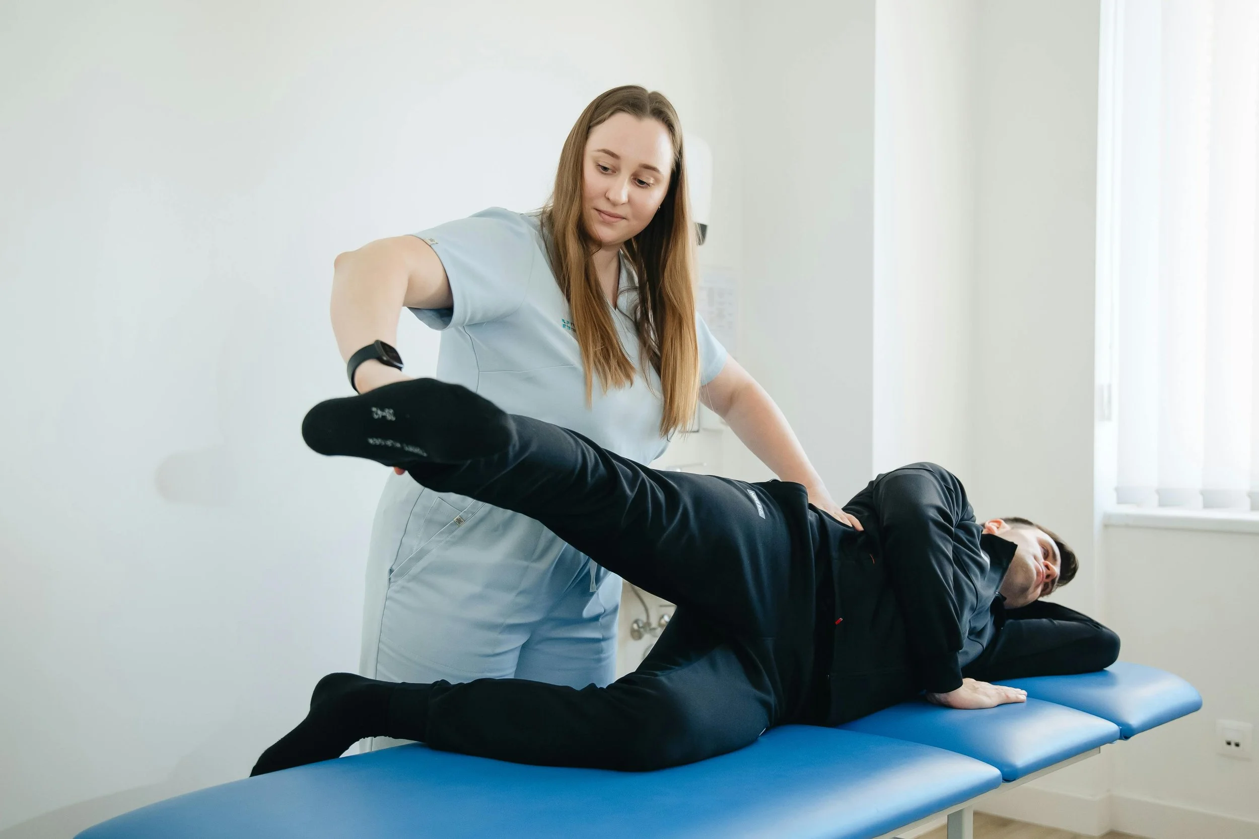 A healthcare professional performs a physical therapy or chiropractic adjustment on a man lying on a blue treatment table, lifting his leg while he lies on his side, in a white, well-lit medical room.
