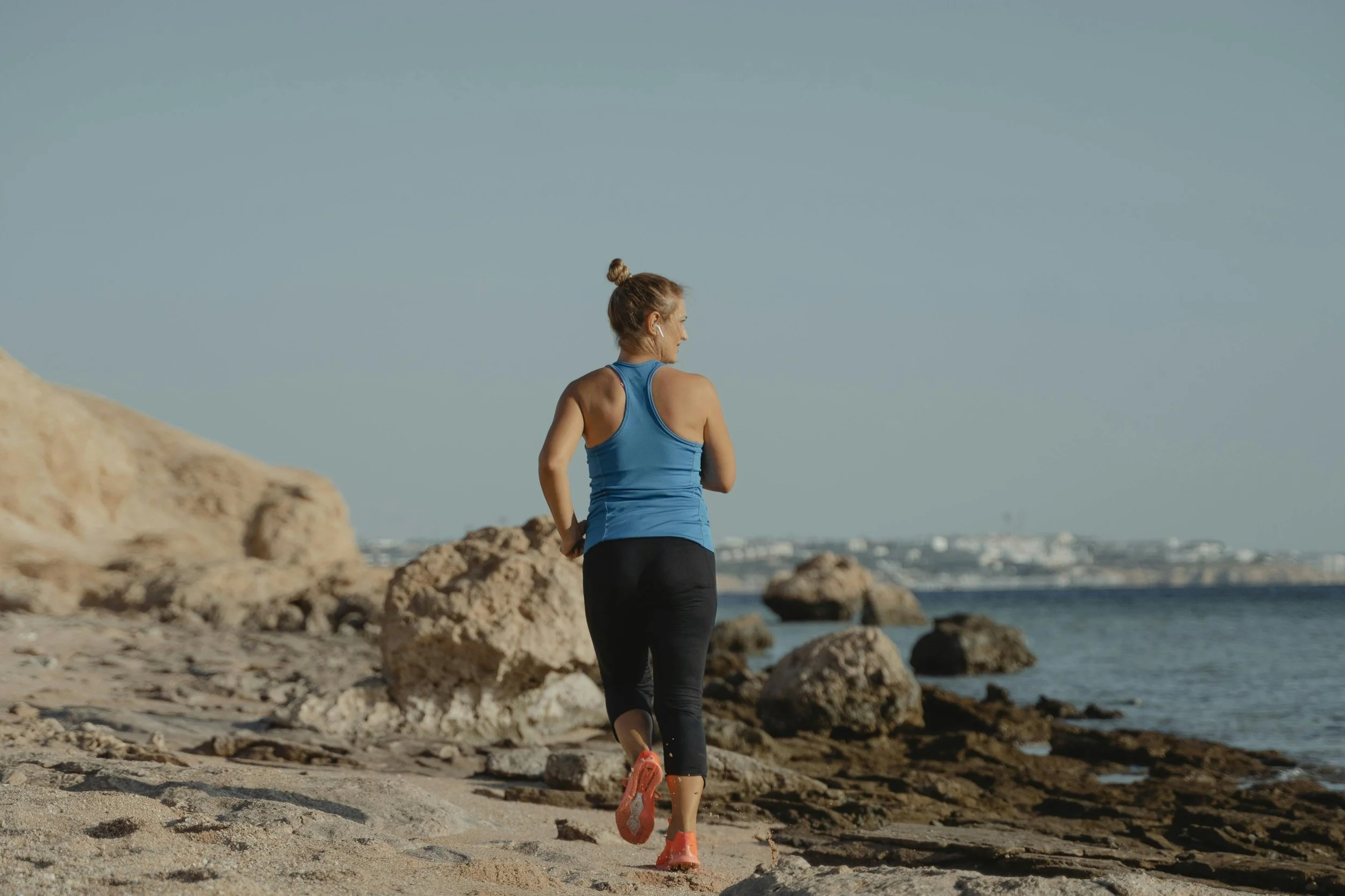 A woman jogging on a rocky beach, wearing a blue tank top, black leggings, and orange running shoes, with the ocean and distant shoreline in the background.