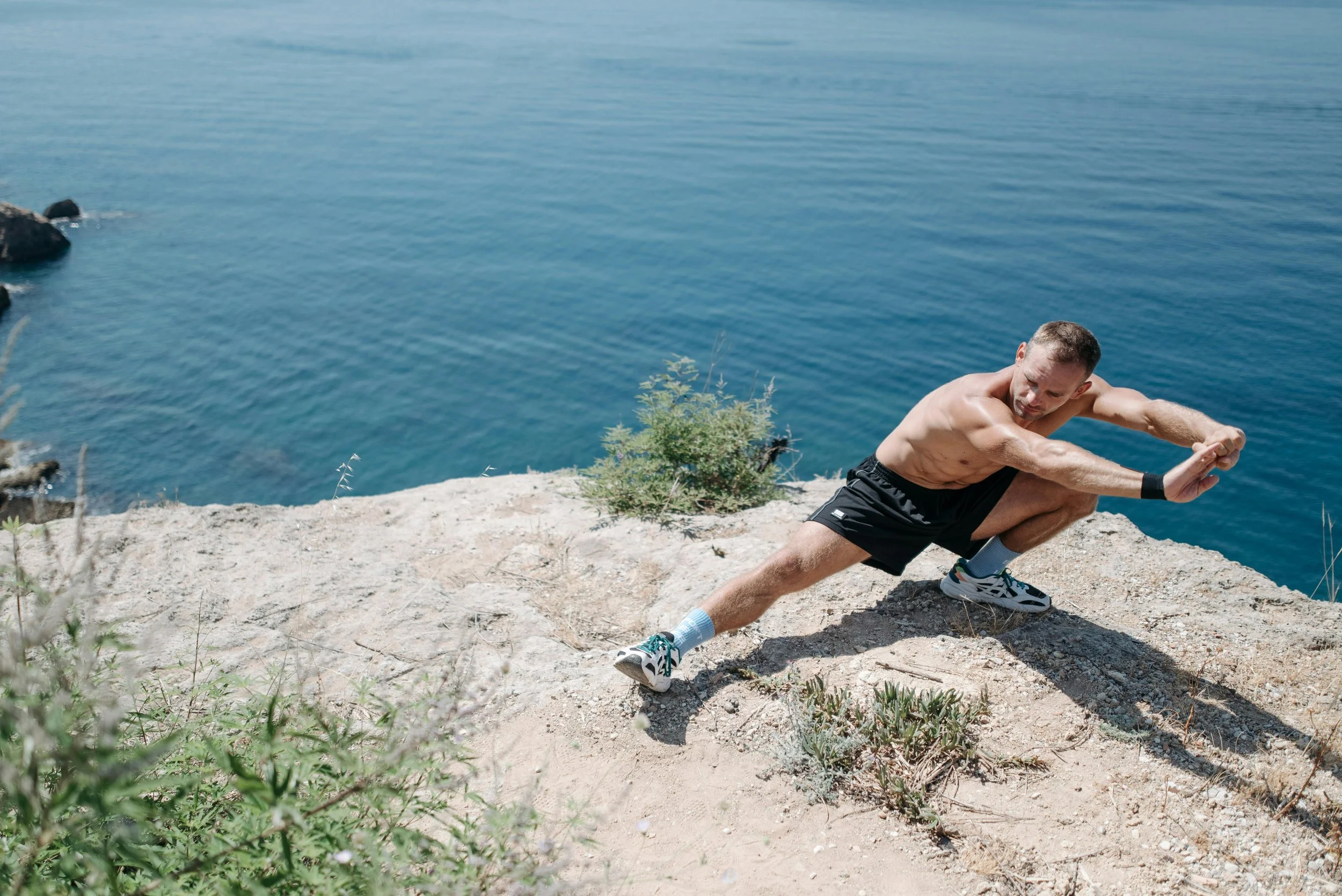 A man doing a wide forward fold stretch on rocky terrain next to a body of water.