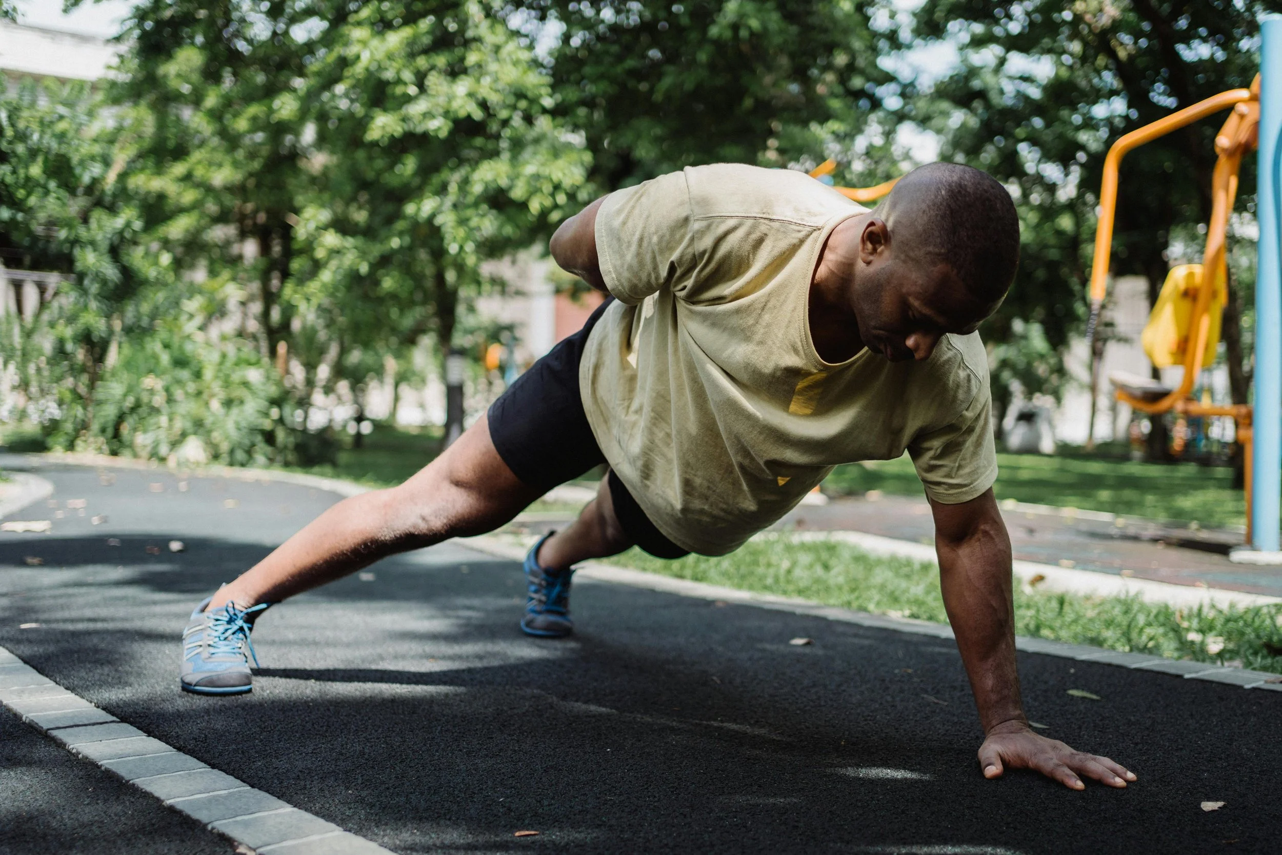 A man doing push-ups outdoors on a black rubber track in a park with trees in the background.