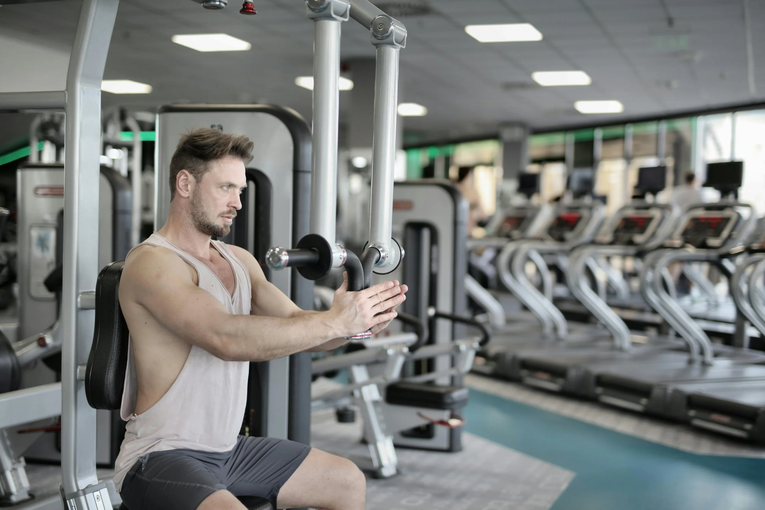 A man in a beige tank top and gray shorts using a chest press machine at the gym, with a row of treadmills in the background.