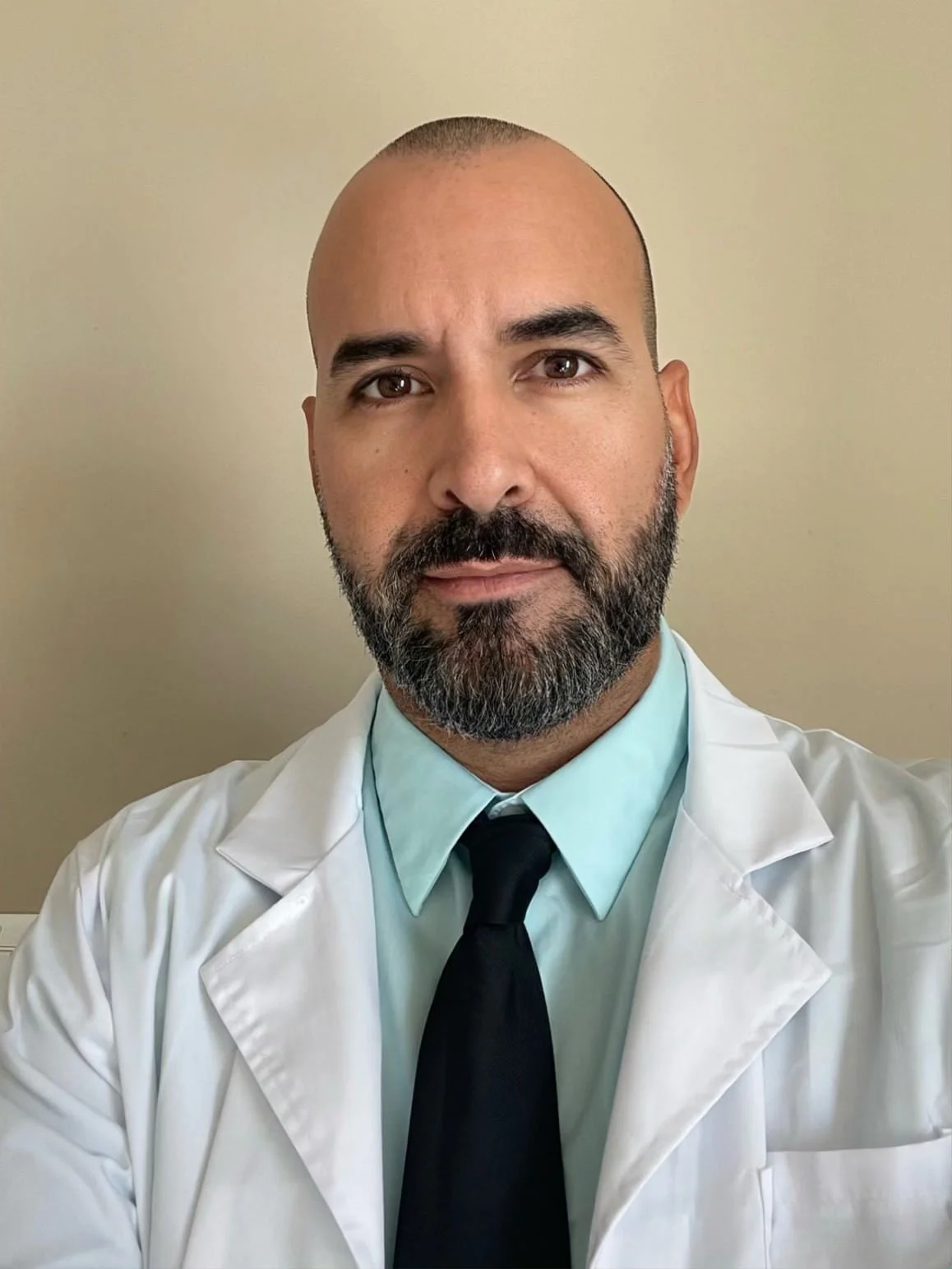 Healthcare professional with a beard and shaved head wearing a white lab coat, light blue shirt, and black tie, standing in front of a plain beige wall.