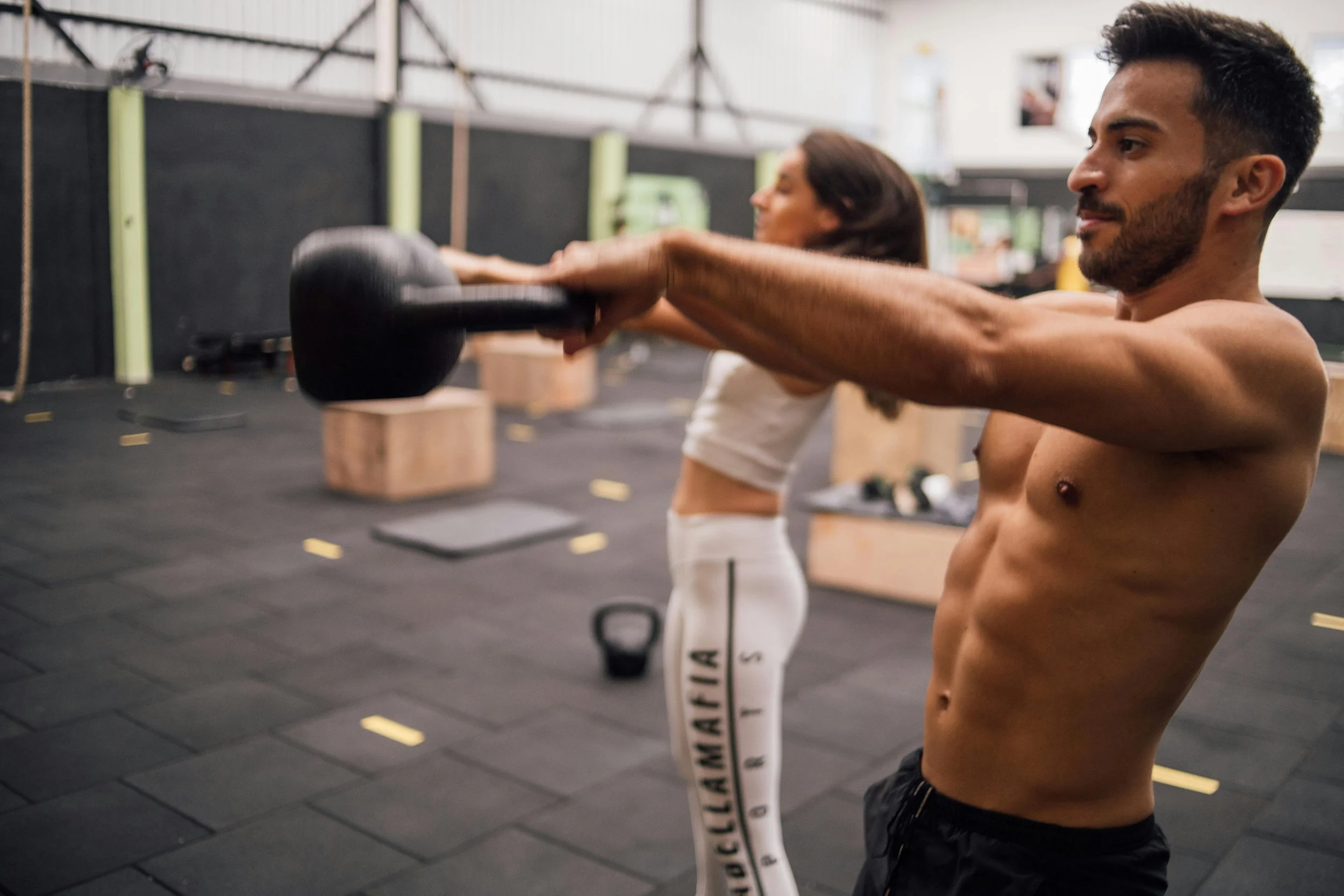 A man and woman are doing a workout in a gym. The man is holding a kettlebell with both hands extended forward. The woman is standing behind him with her arms stretched out, facing away. They are both dressed in athletic wear.