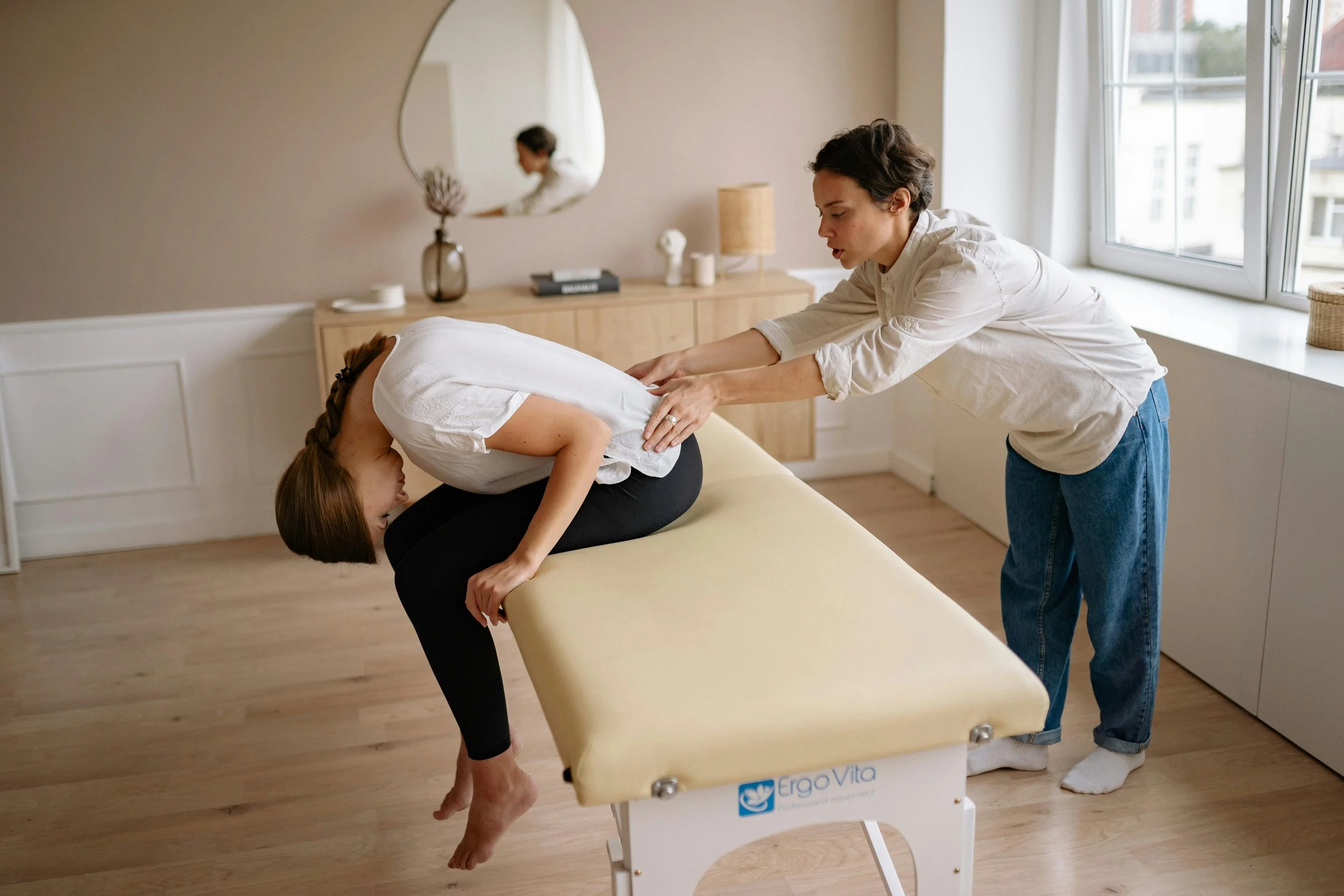 A woman is receiving a massage or physical therapy from a therapist in a clinical setting while sitting on a therapy table. The woman is bent over with her head down, and the therapist is applying pressure to her back.