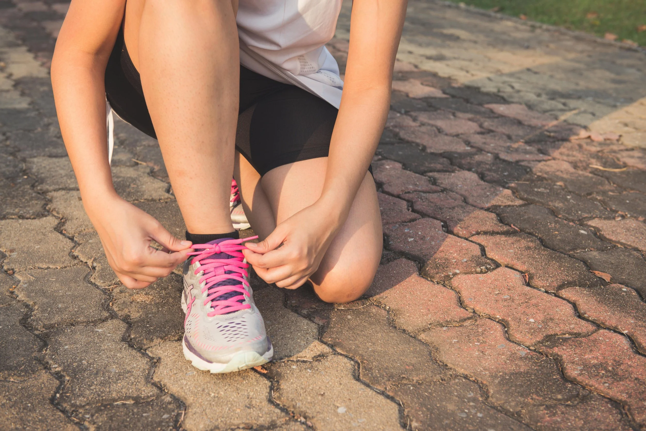 A person kneeling on a paved outdoor path, tying the pink laces of their running shoe, wearing black shorts and a white shirt.