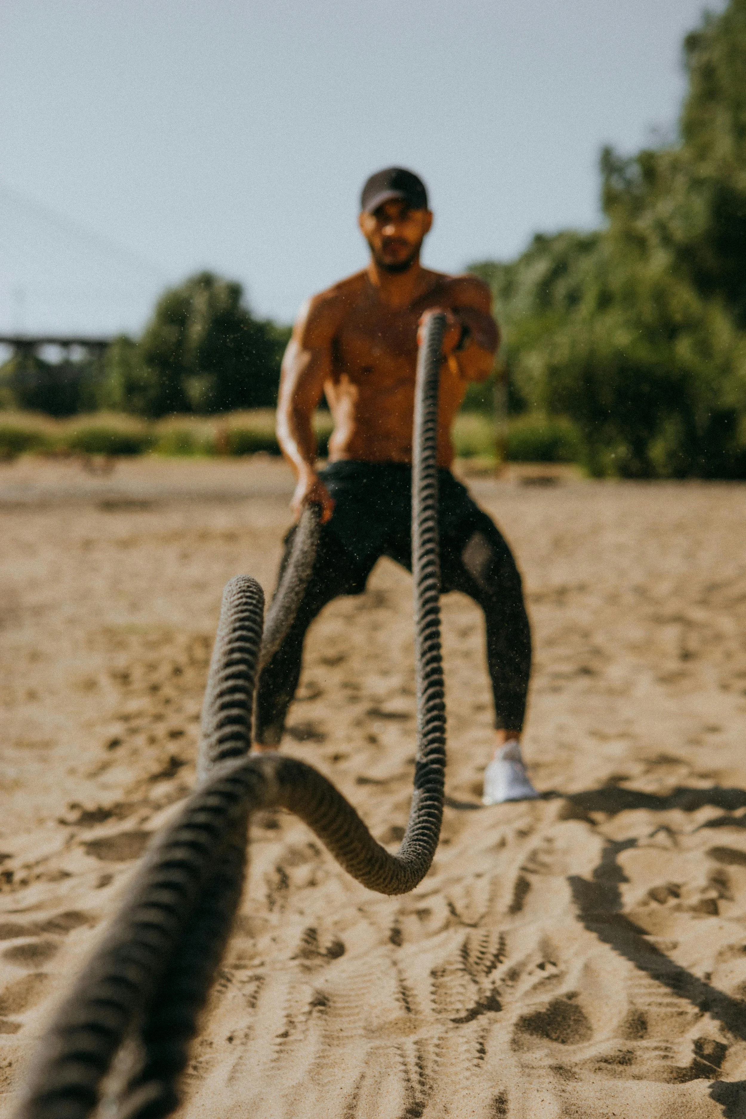 A  man in black workout pants doing battle rope workout on a sandy outdoor surface.