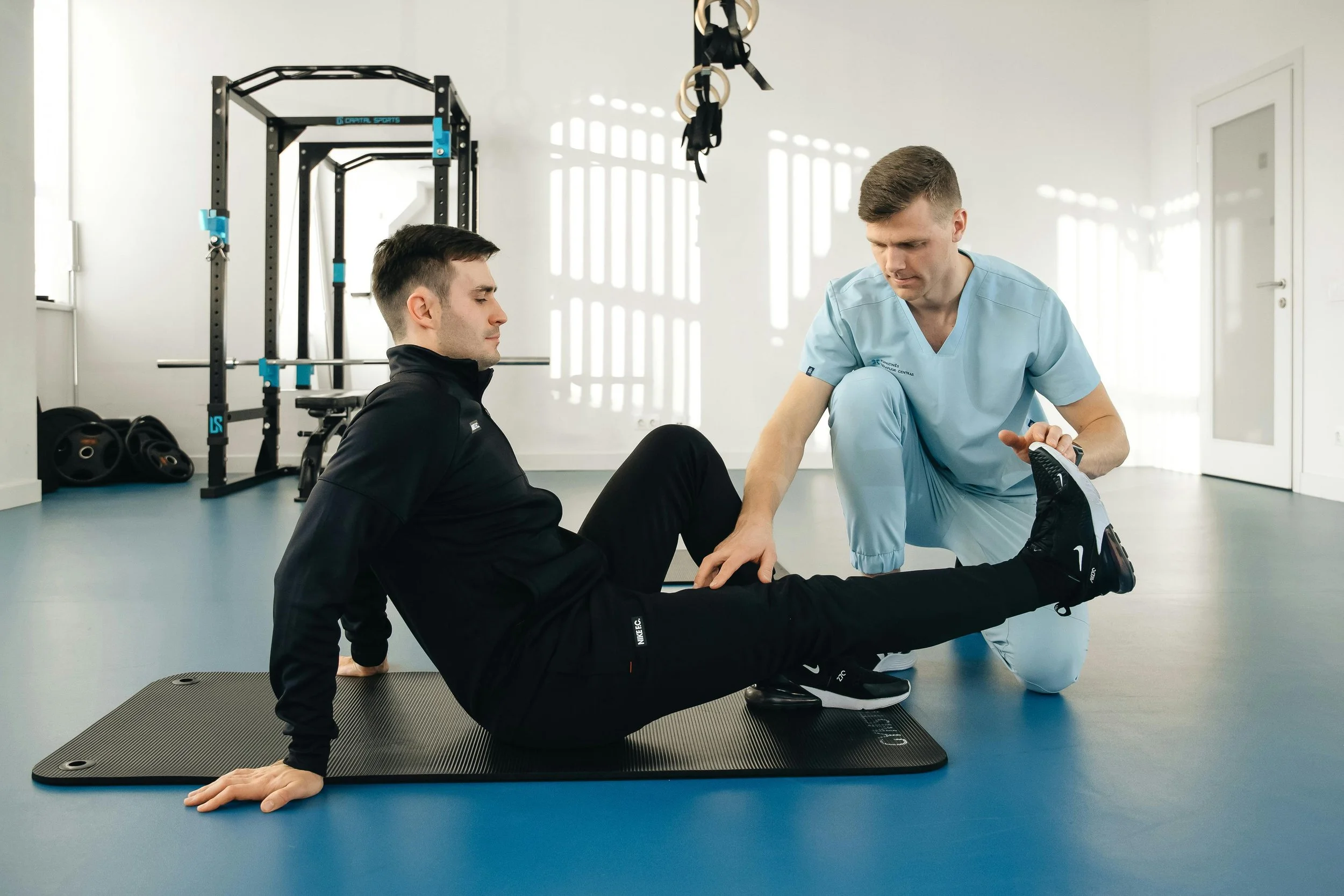 A physiotherapist assisting a male patient with a leg stretch exercise in a fitness center.