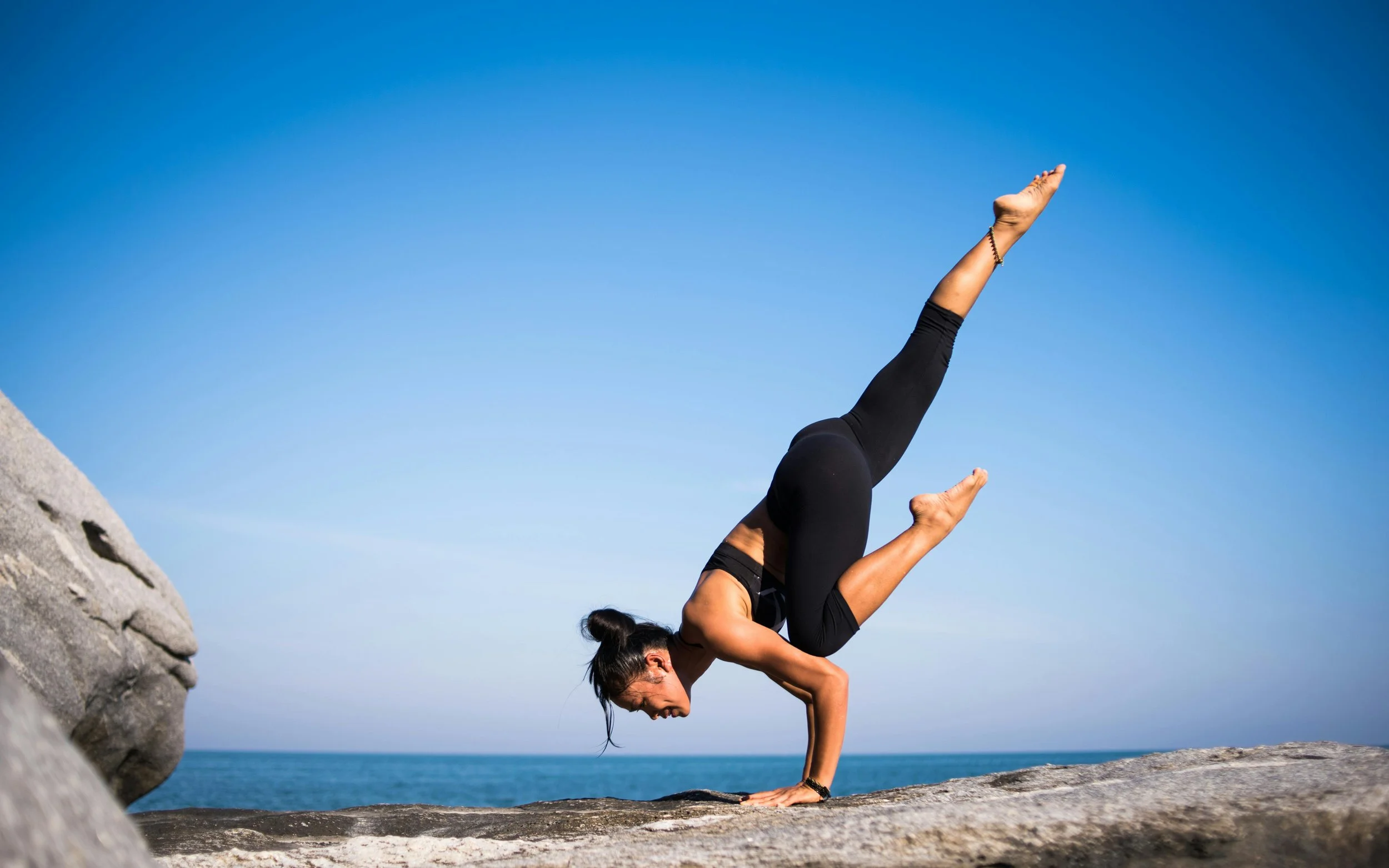A woman practicing yoga outdoors on a rocky surface near the ocean, performing a forearm stand with legs extended upward against a clear blue sky.