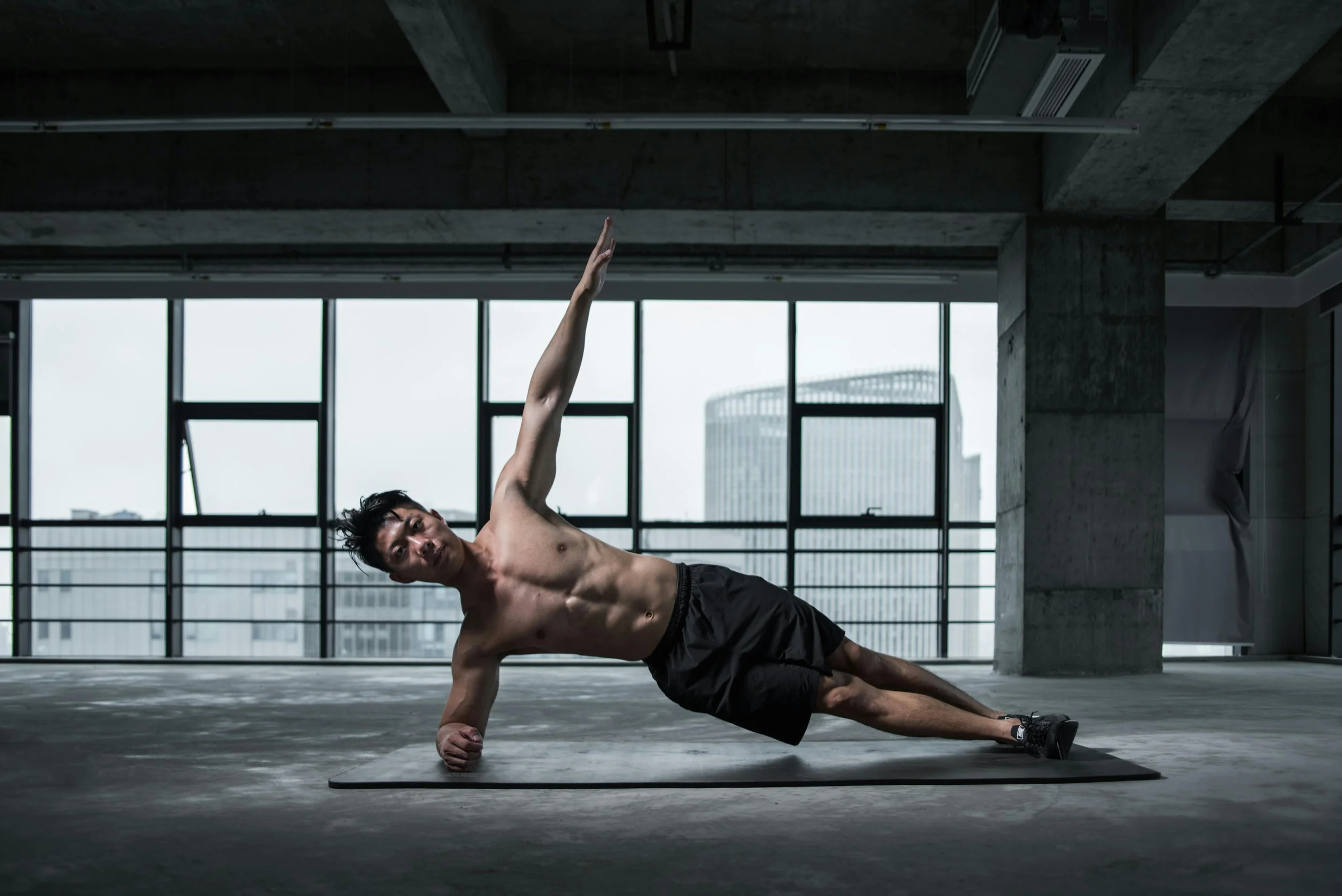A shirtless man in black shorts and sneakers doing a side plank exercise on a yoga mat in an industrial-style room with large windows and city buildings outside.