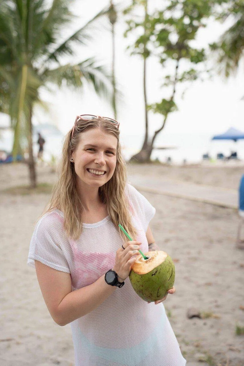 woman is in a bathing suit on a beach drinking water out of a coconut and smiling at the camera