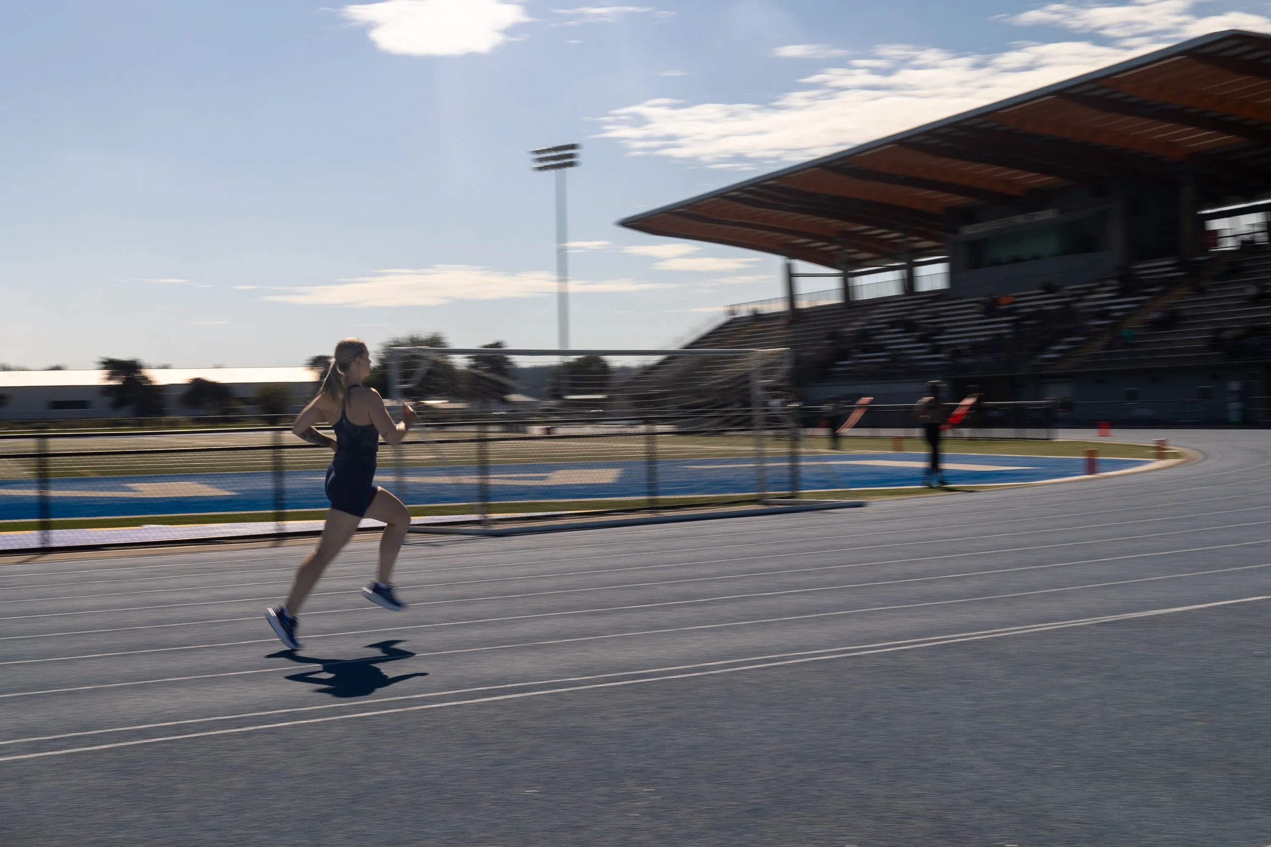 Female in black athletic wear running on an outdoor track at a sports stadium.