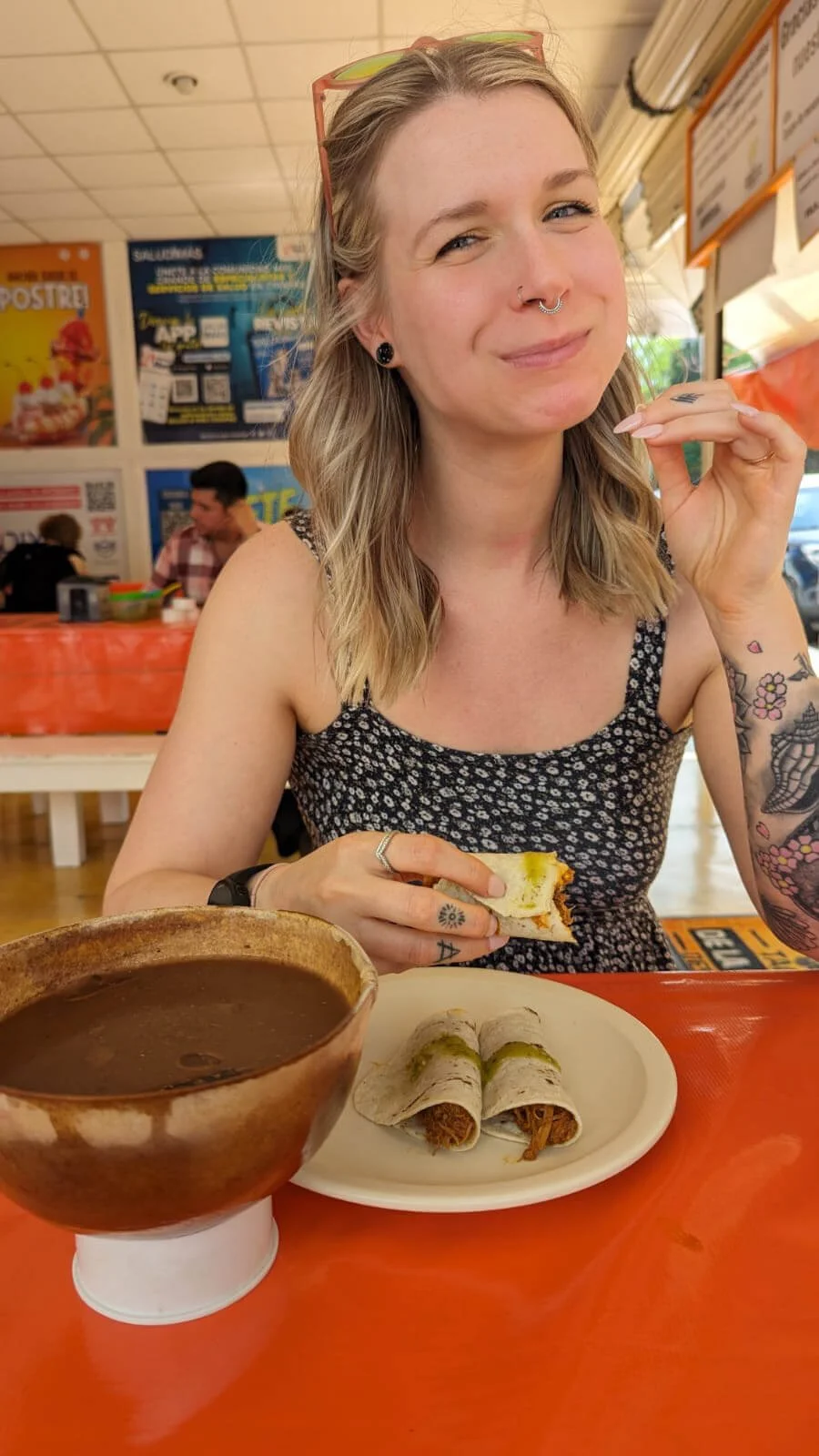 woman with blonde hair is smiling at camera while eating tacos in a brightly coloured restaurant